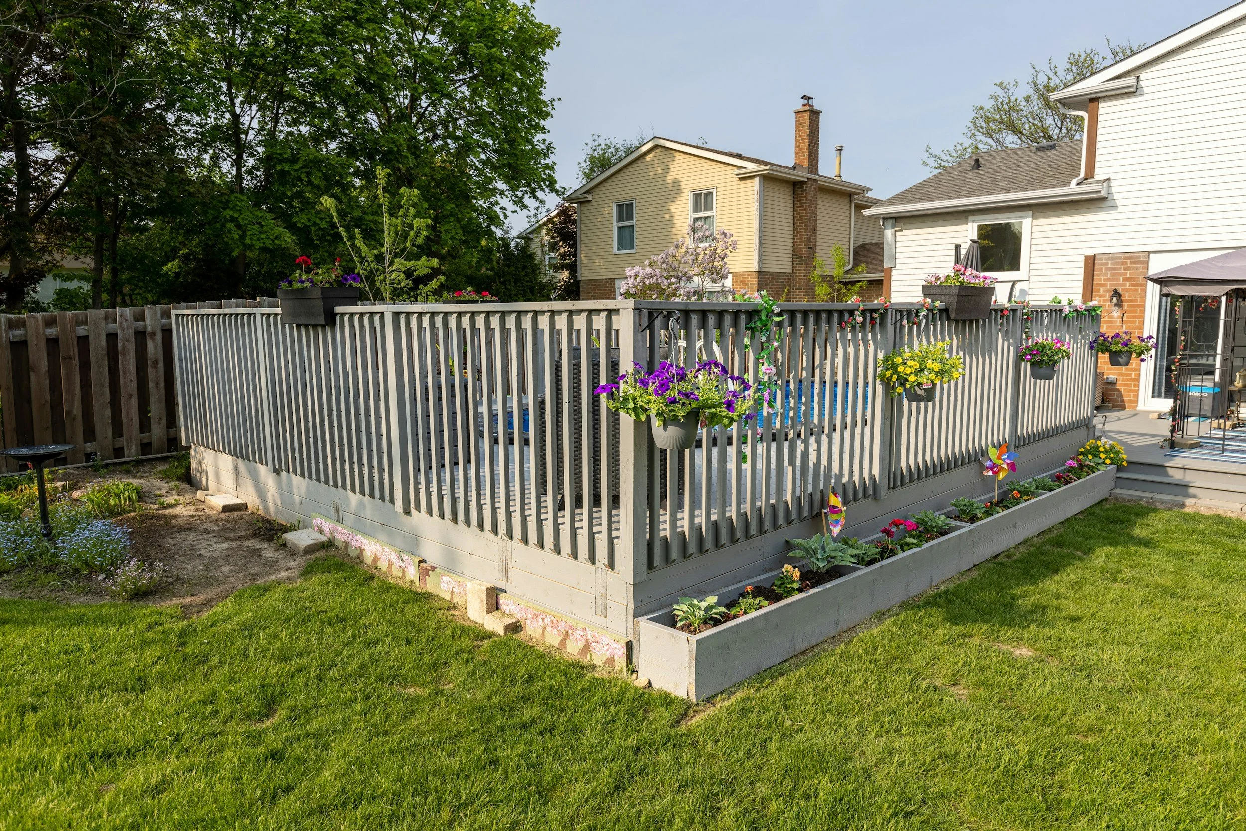 Backyard with a wooden deck, potted flowers, a garden bed with colorful flowers, and neighboring houses in the background.