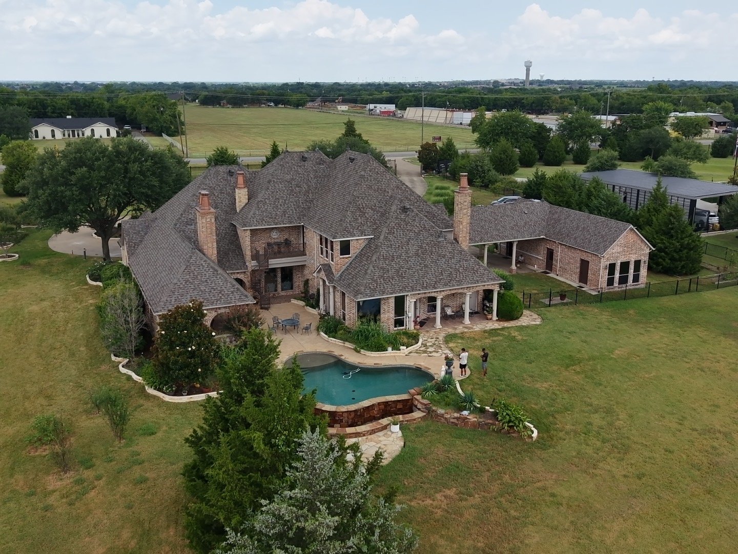 Aerial view of a large brick house with a swimming pool, surrounded by green lawn and trees, in a rural area with fields and other buildings in the background.