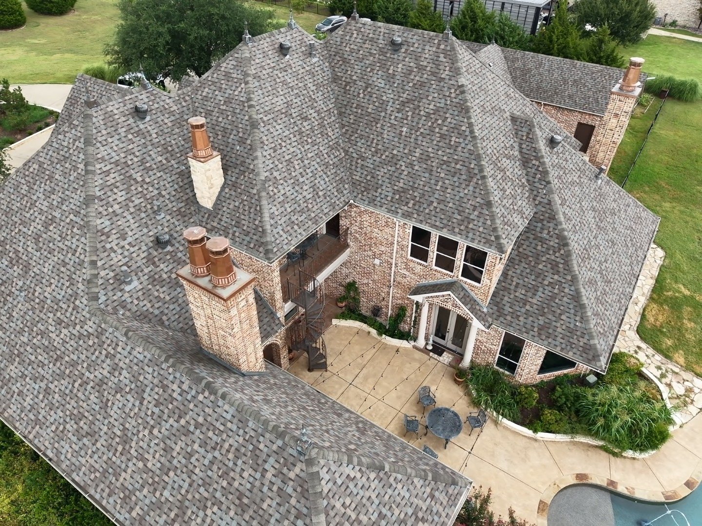 Aerial view of a large brick house with a complex gray shingle roof, multiple chimneys, and a backyard patio with outdoor furniture and landscaping.
