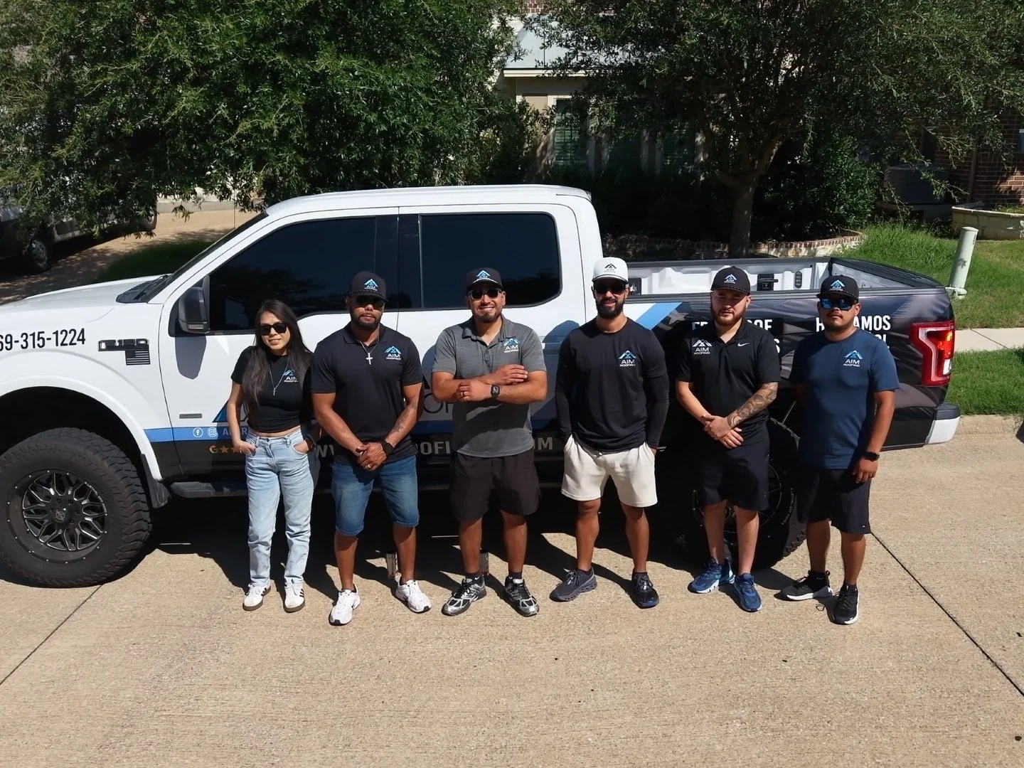 Six people standing in front of a white and dark blue police or security pickup truck, outdoors on a sunny day, with trees and a house in the background. They are wearing casual or team uniforms, some with caps and sunglasses.