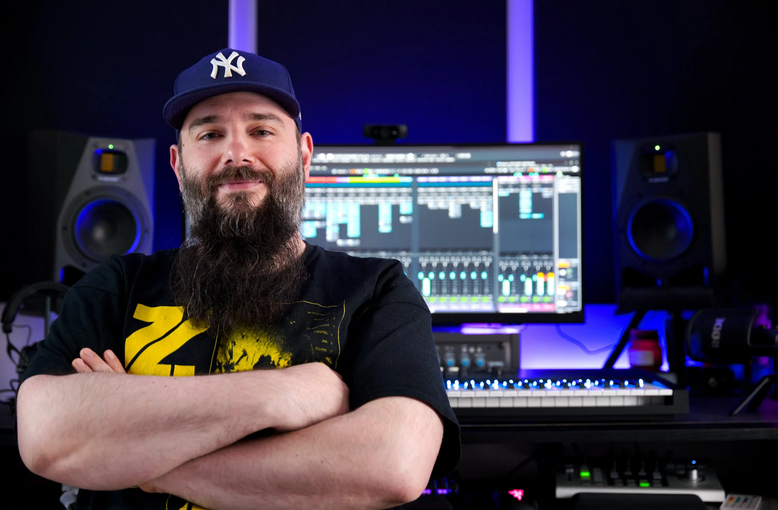 A bearded man with a cap in front of a music production setup with two large monitors displaying digital audio workstation software, speakers, and various audio equipment.