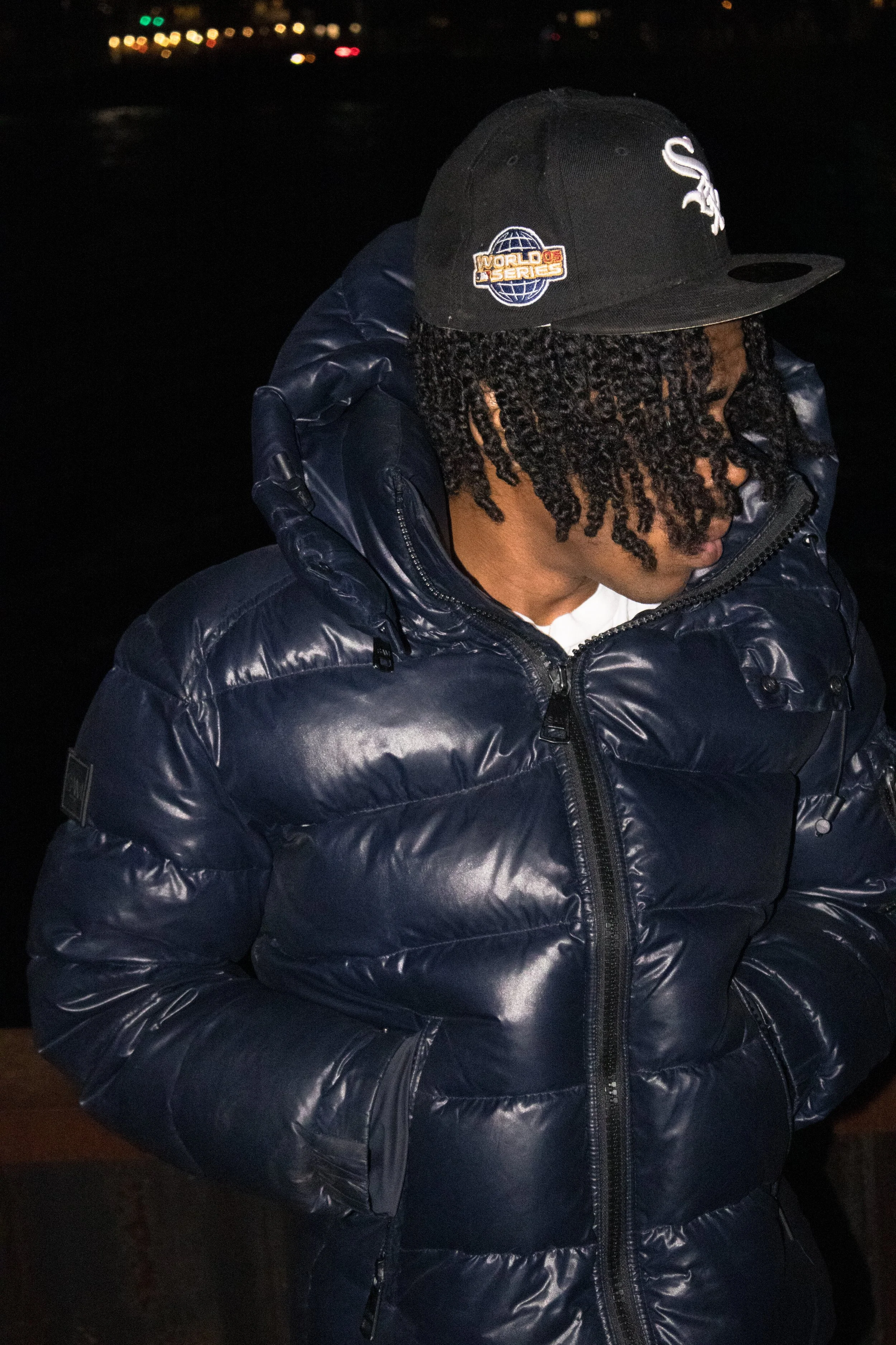 A person with dreadlocks wearing a dark blue puffer jacket and a black baseball cap with a sports logo, looking down outside at night.