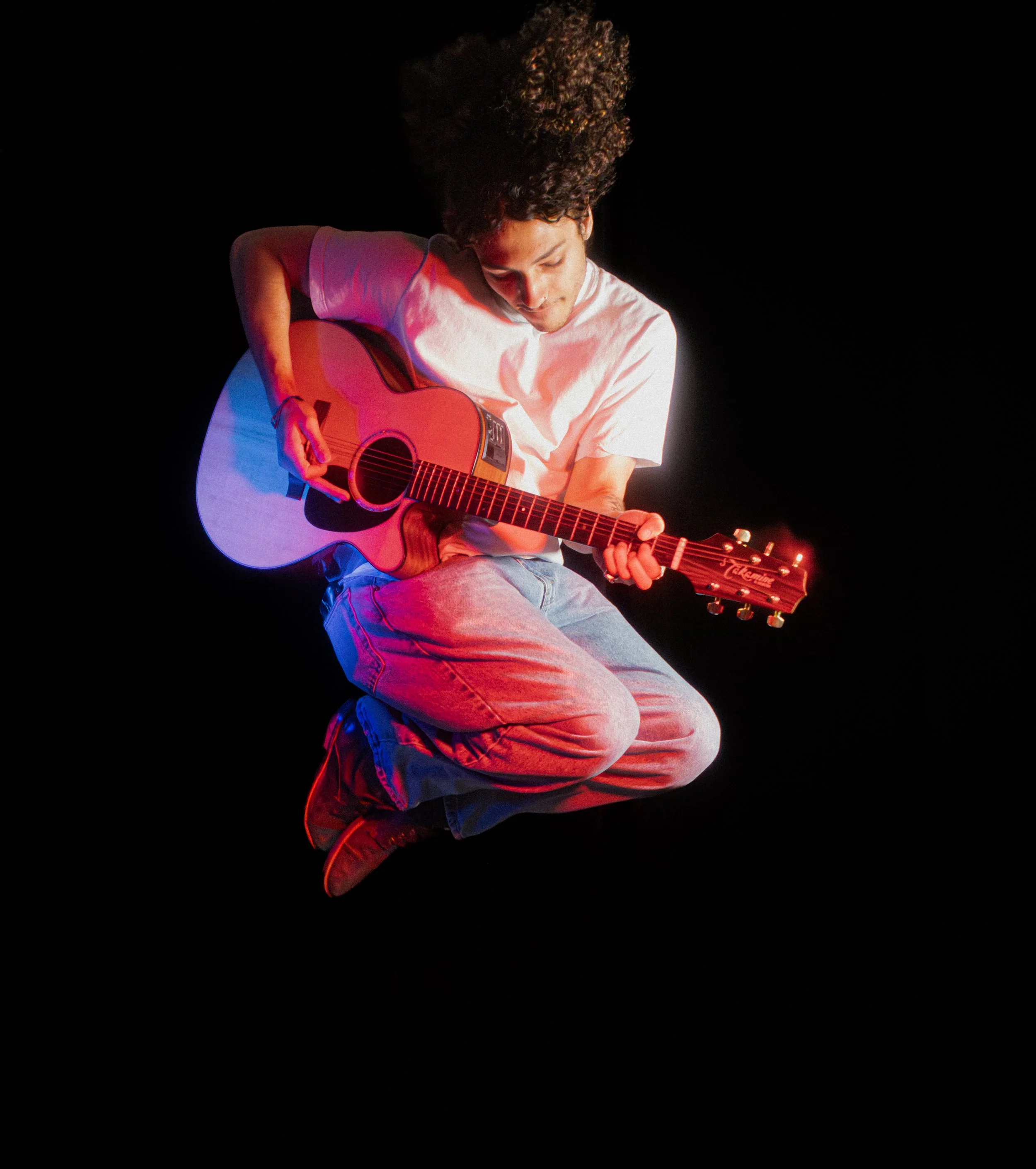 A person with curly hair wearing a white t-shirt and jeans, sitting in a cross-legged position, playing an acoustic guitar against a black background.