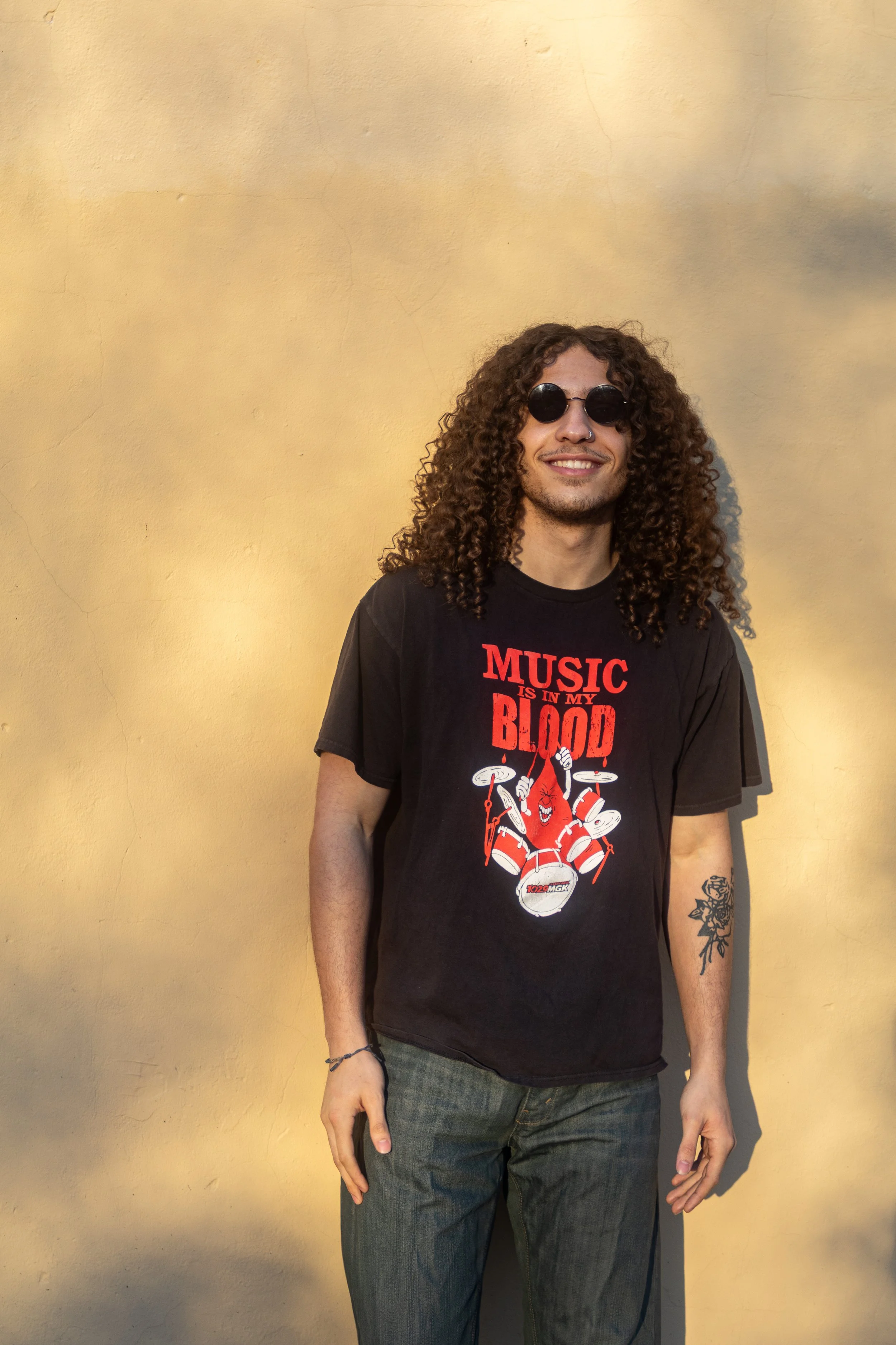 A man with curly hair, sunglasses, and a nose ring, standing against a yellow wall, wearing a black t-shirt with red and white text and graphics.