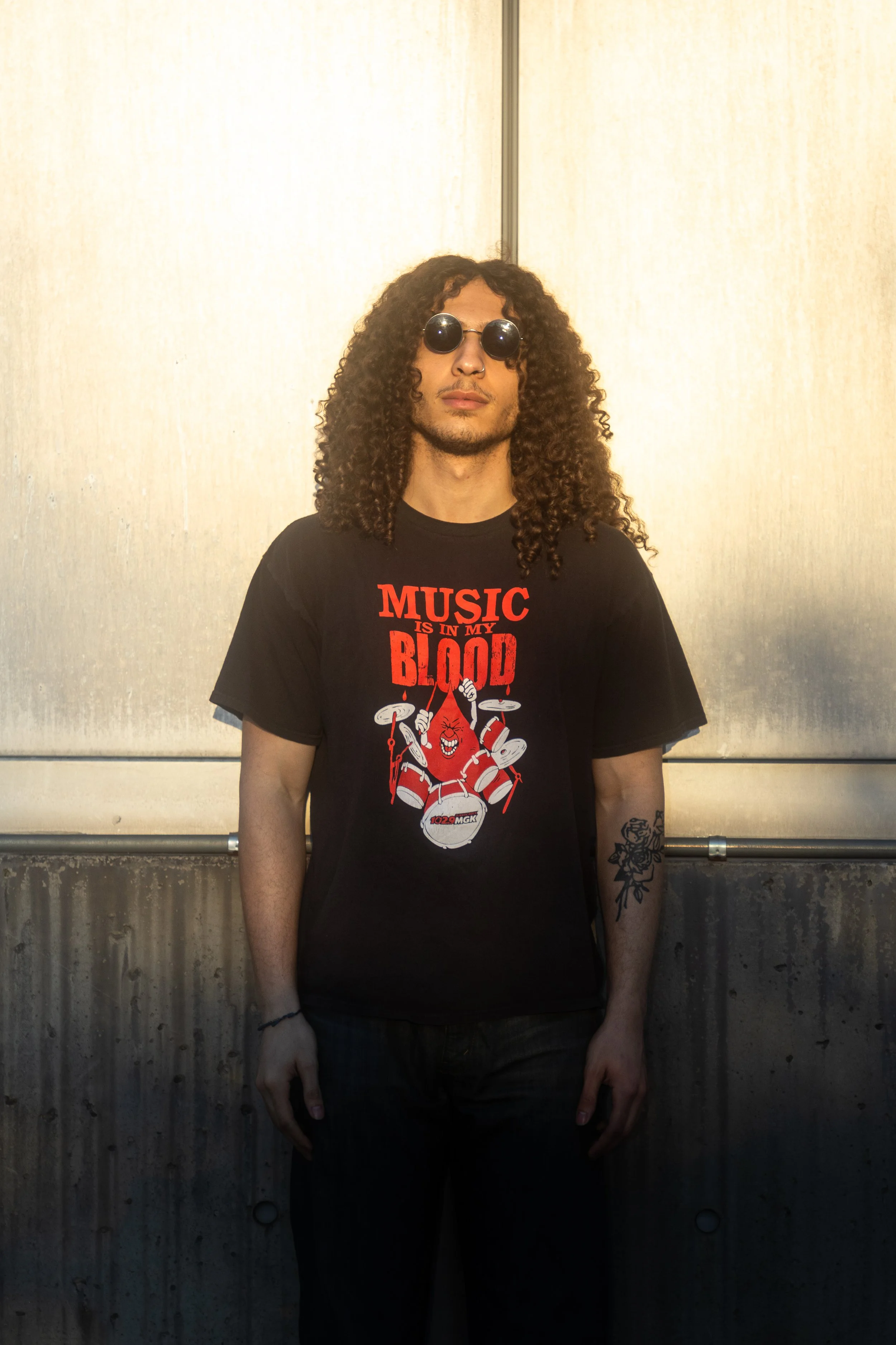 A young man with curly hair wearing sunglasses and a black T-shirt with a graphic of a blood-dripping cartoon character and the text 'Music is in my blood', standing against a metallic background.