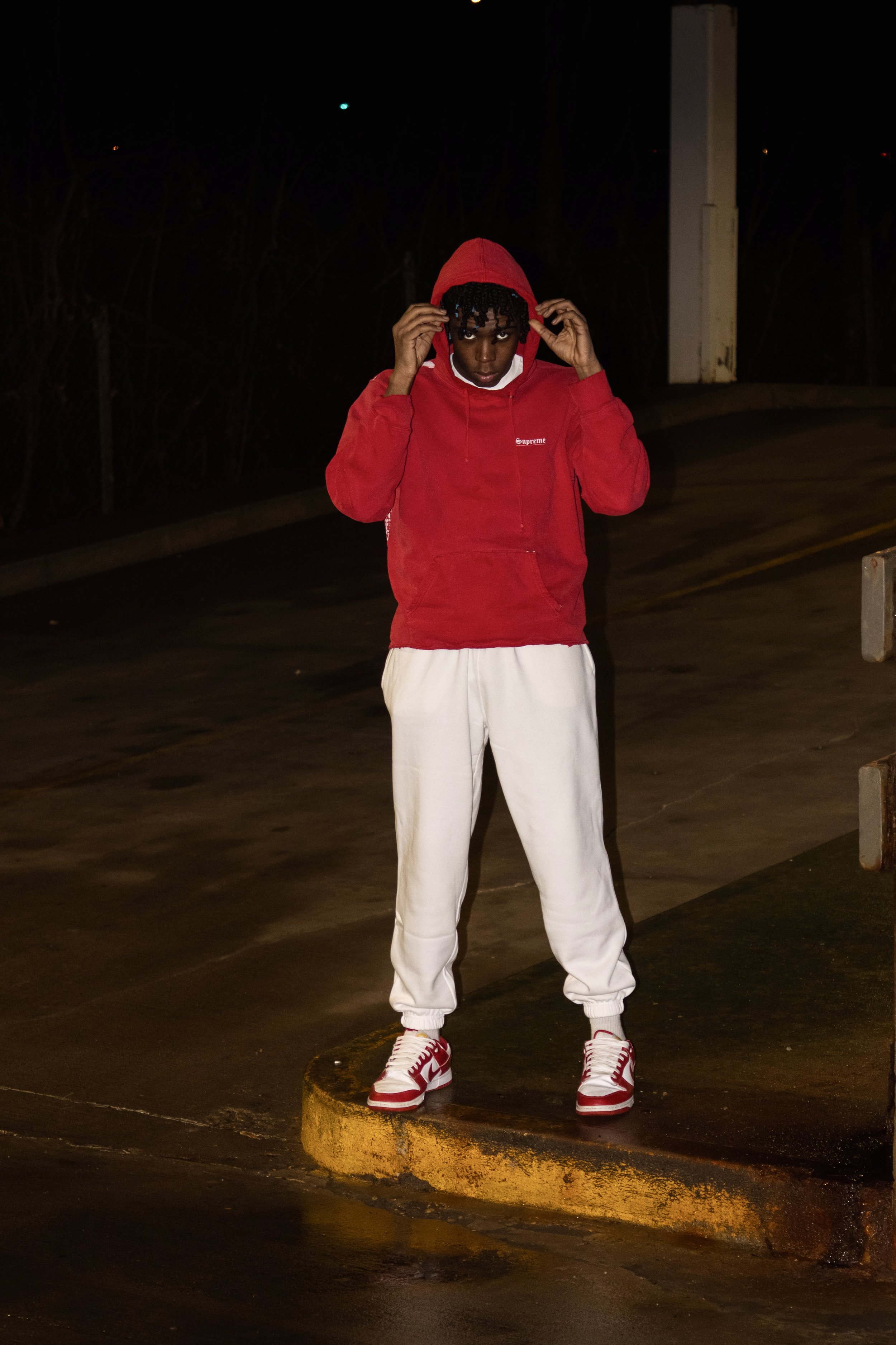 A young man wearing a red hoodie, white sweatpants, and red sneakers standing on a parking curb at night, adjusting his hood.
