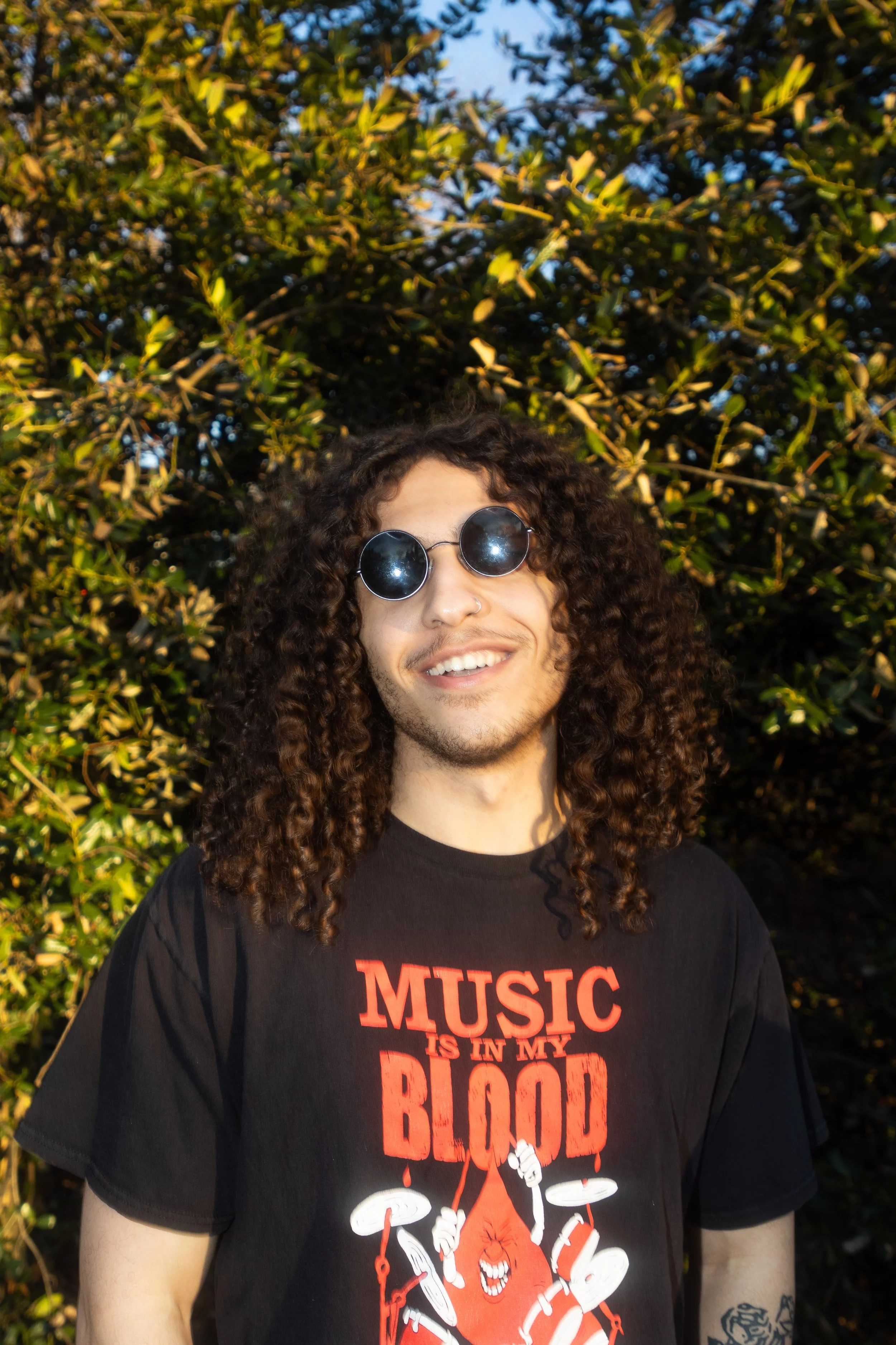 A young man with curly hair wearing round sunglasses and a black T-shirt with red and white text and graphics, standing outdoors in front of green foliage.