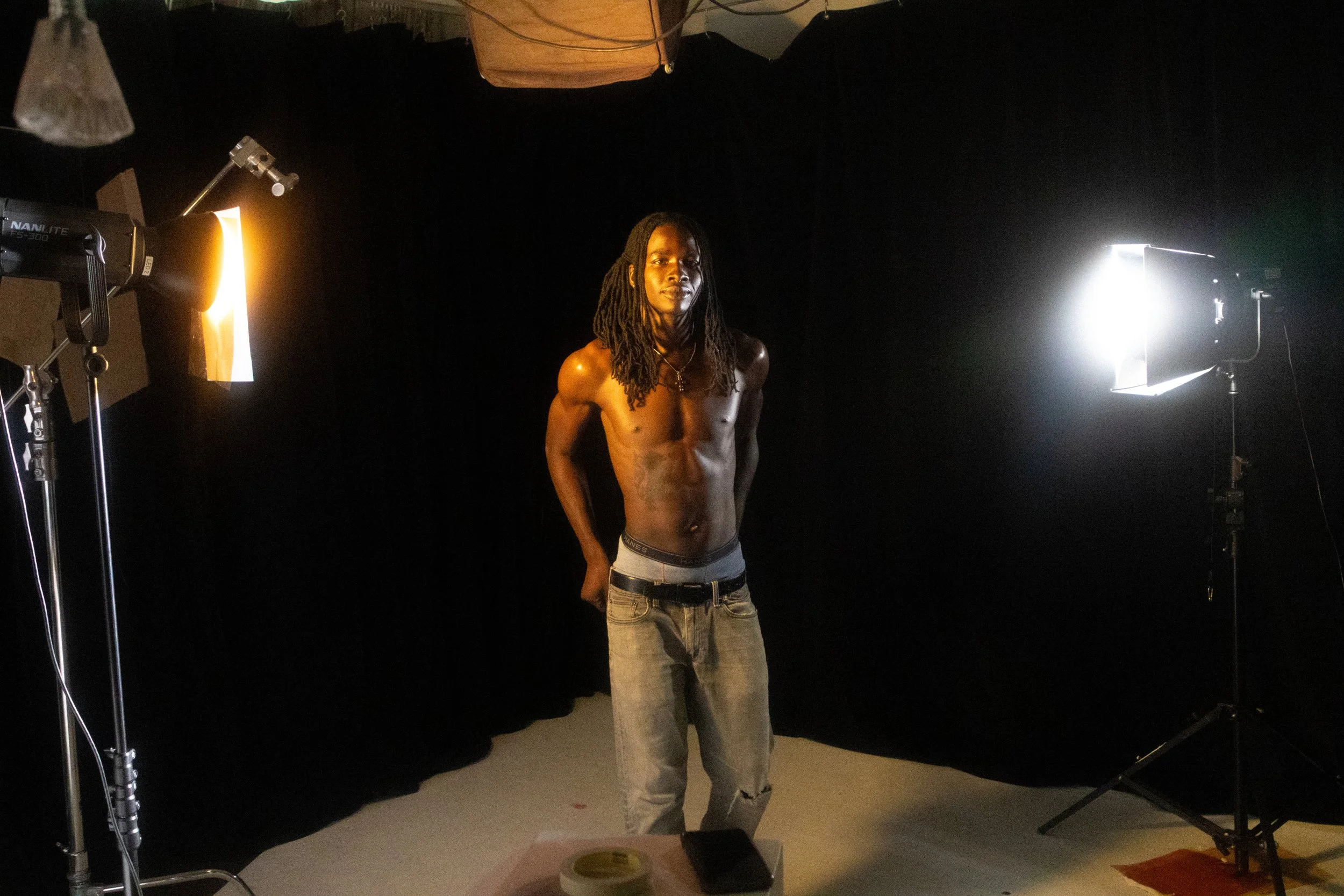 A shirtless man with long dreadlocks stands in a photography studio with black background and professional lighting equipment.