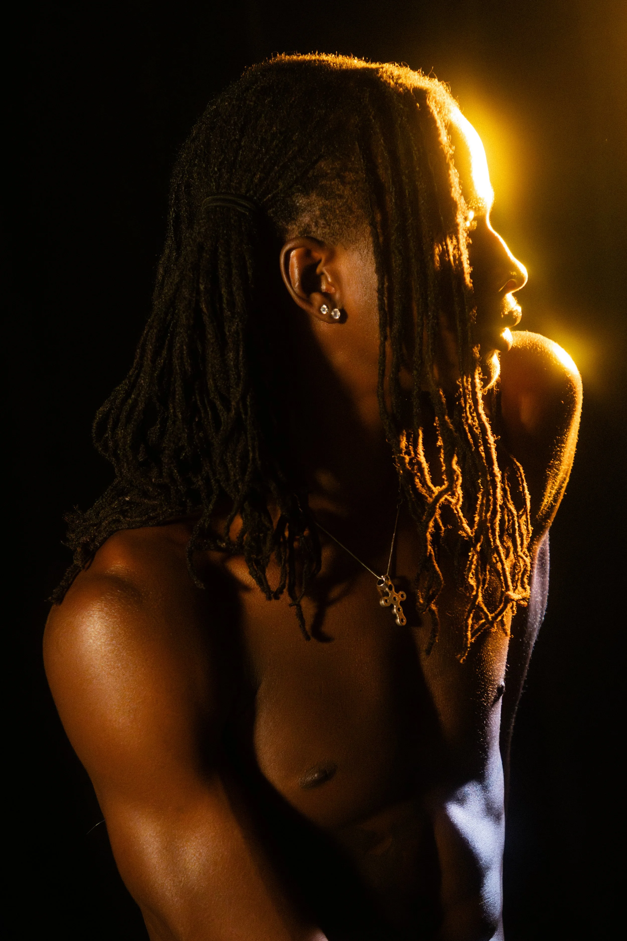 A side profile of a shirtless person with dreadlocks, earrings, and a necklace, illuminated by warm backlighting against a dark background.