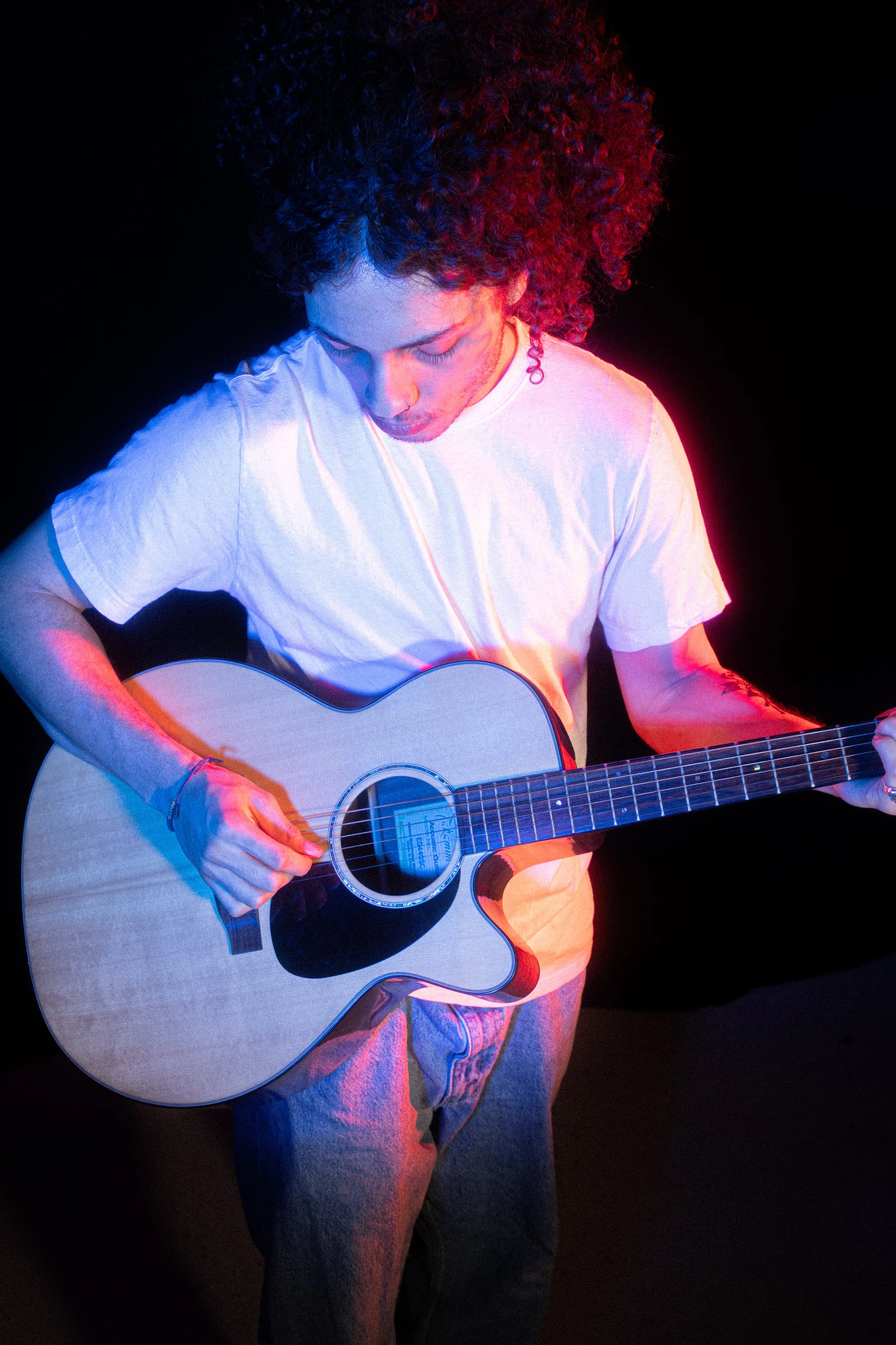 Person with curly hair playing an acoustic guitar on stage, illuminated by colorful stage lights.