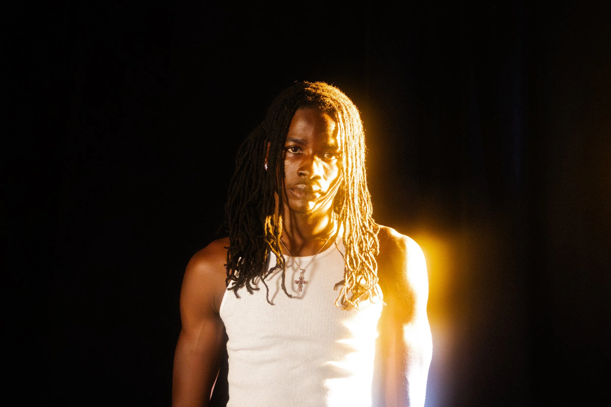 A person with long dreadlocks wearing a white tank top, captured in dramatic lighting with a dark background.