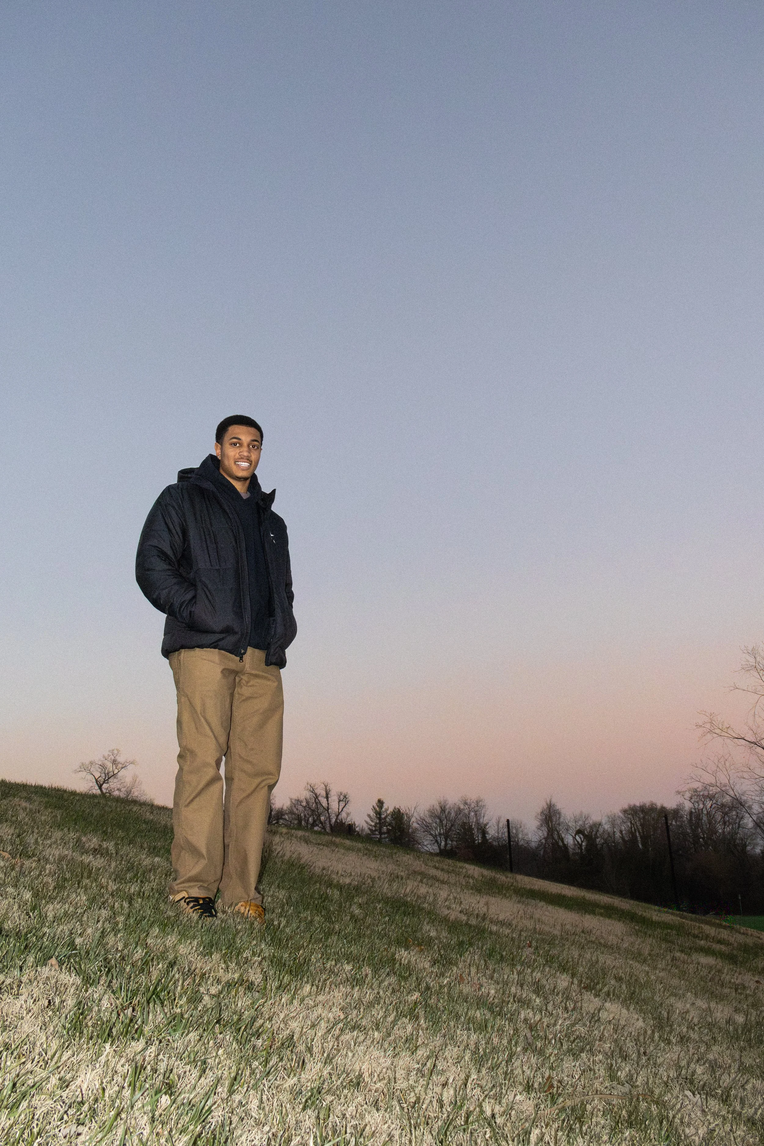 A young man standing outdoors on a grassy hill at dusk, wearing a black jacket, khaki pants, and brown shoes, smiling at the camera with trees in the background.