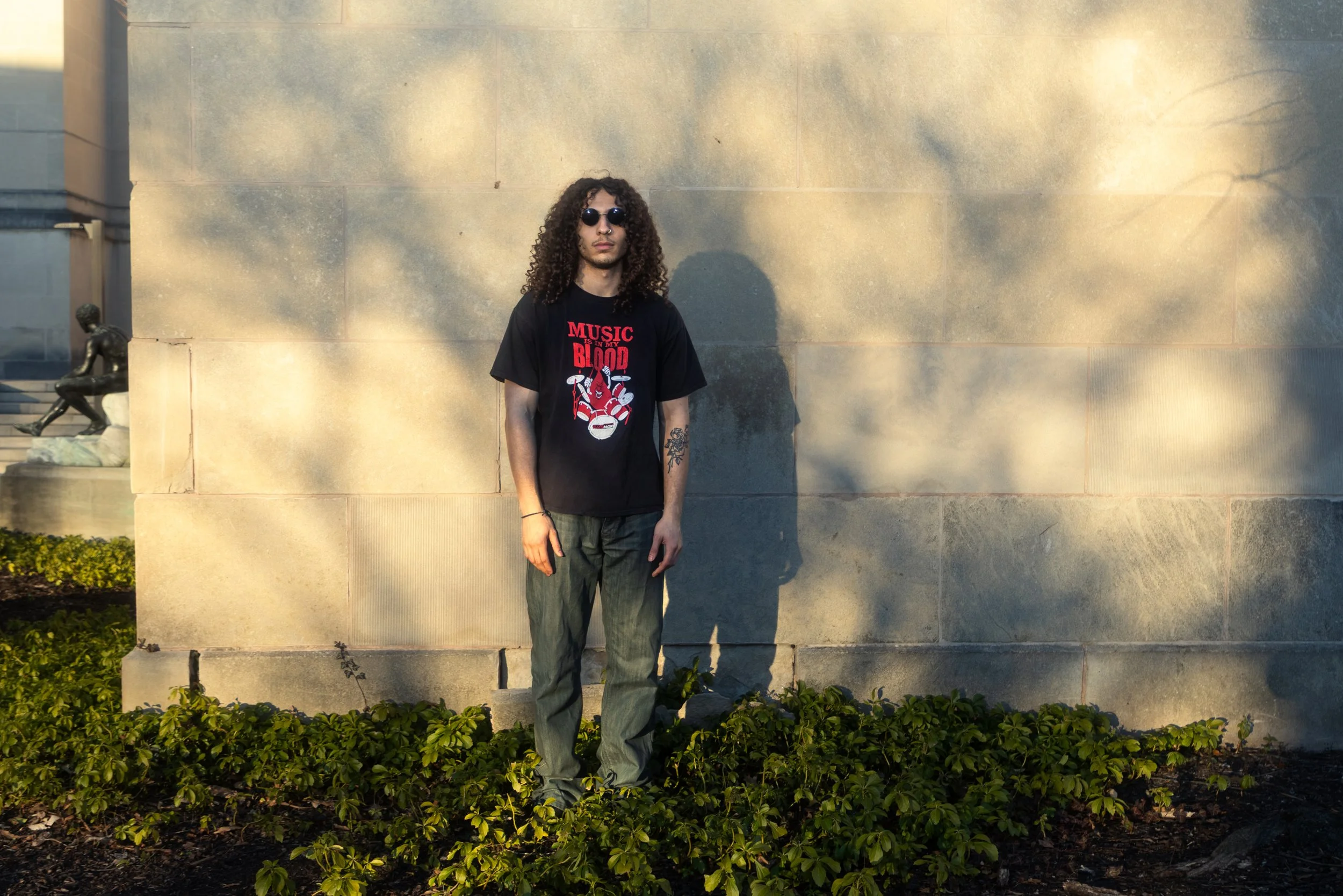 A young man with long curly hair and sunglasses standing against a stone wall, with sunlight casting his shadow on the wall. He is wearing a black T-shirt with a graphic design and text, and gray pants, surrounded by green bushes at his feet.