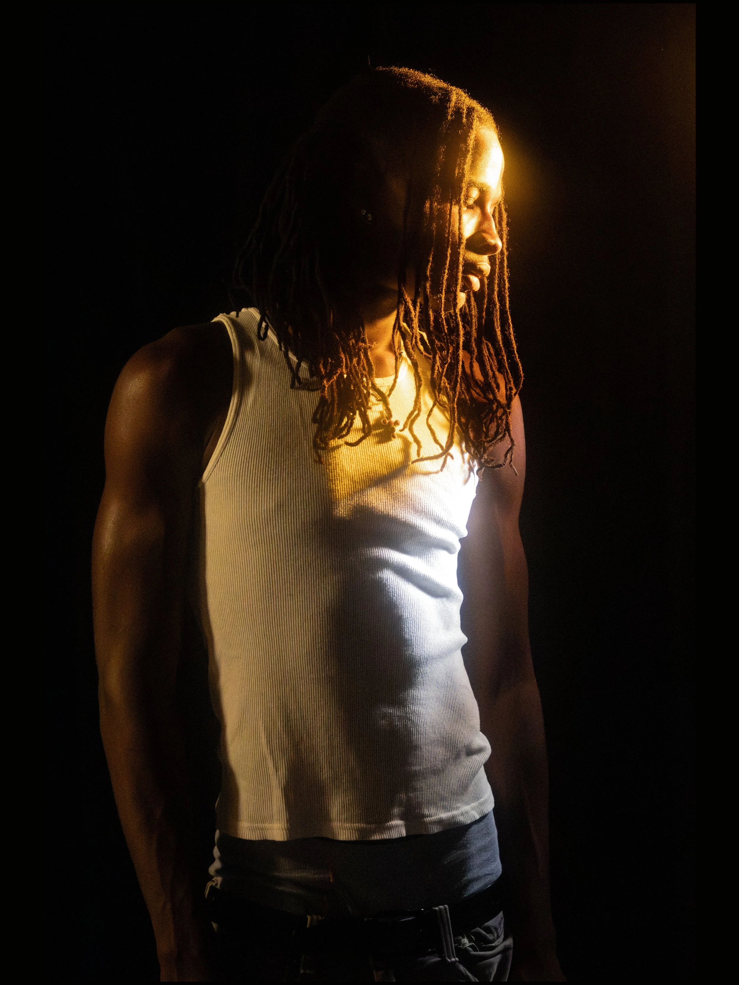 A man with dreadlocks, wearing a white sleeveless shirt, standing in darkness with a warm light illuminating his face and upper body.
