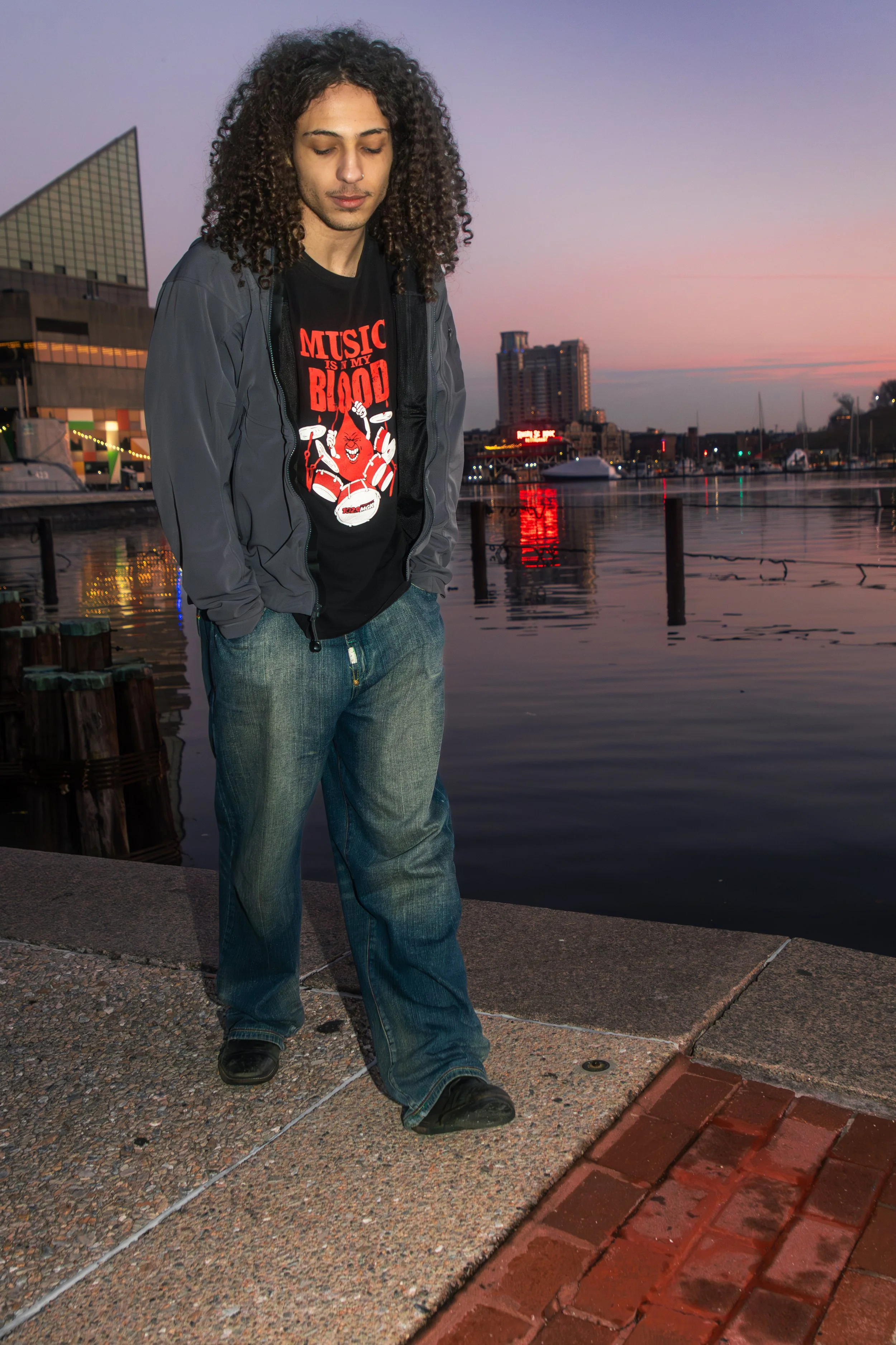 Young man with curly hair standing on a waterfront promenade during sunset, wearing a black graphic t-shirt that says 'Music is my Blood,' a gray jacket, and baggy jeans.