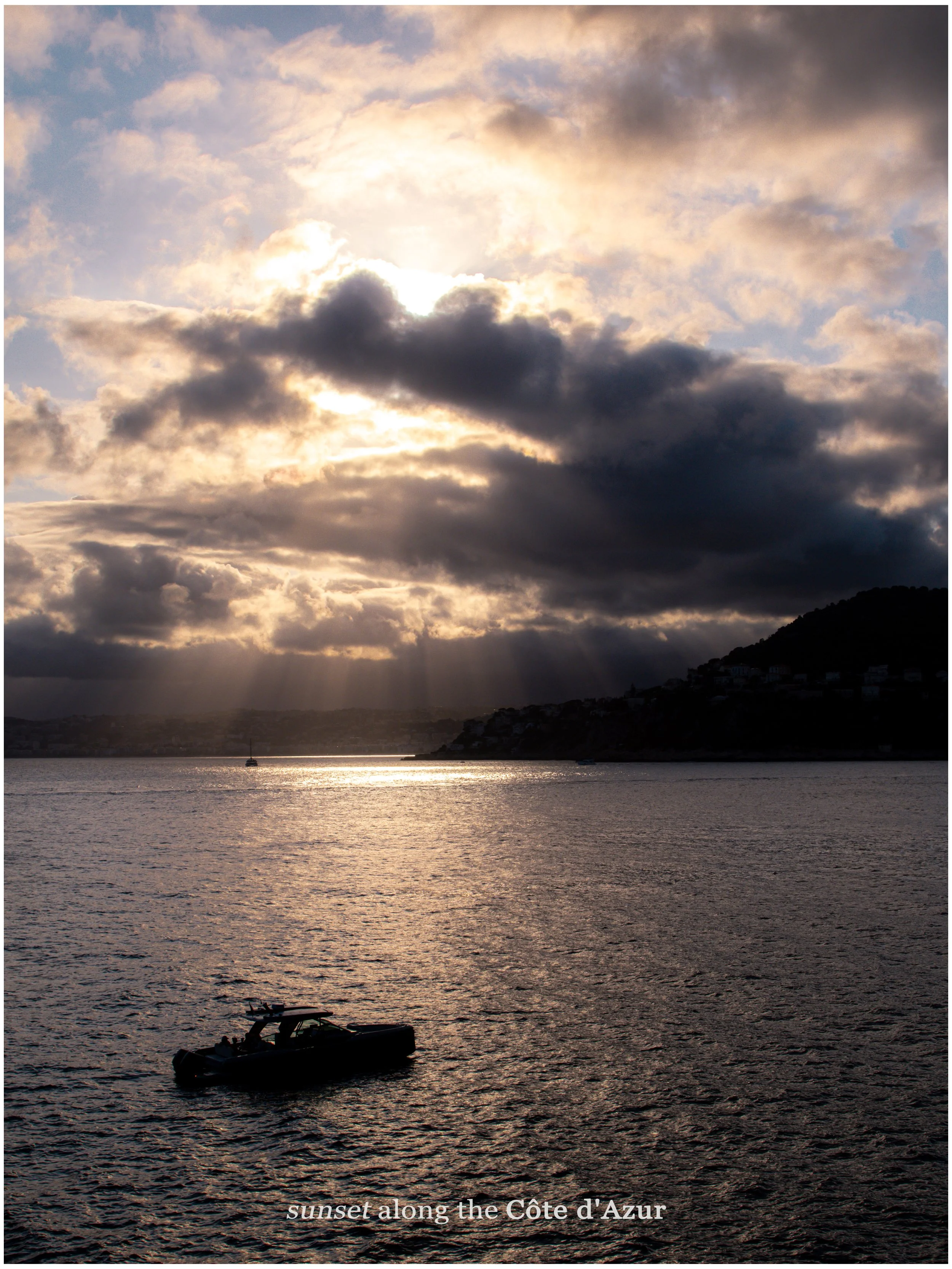Sunset over the water with a boat in the foreground and a cloudy sky with sun rays peeking through, along the Côte d'Azur.