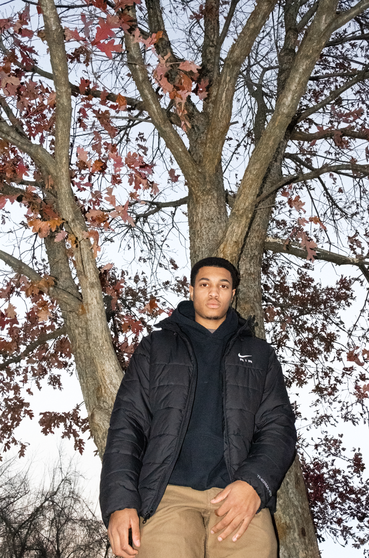A young man standing outdoors in front of a large tree with brown and reddish leaves. The sky is cloudy, and the man is wearing a black jacket and tan pants.