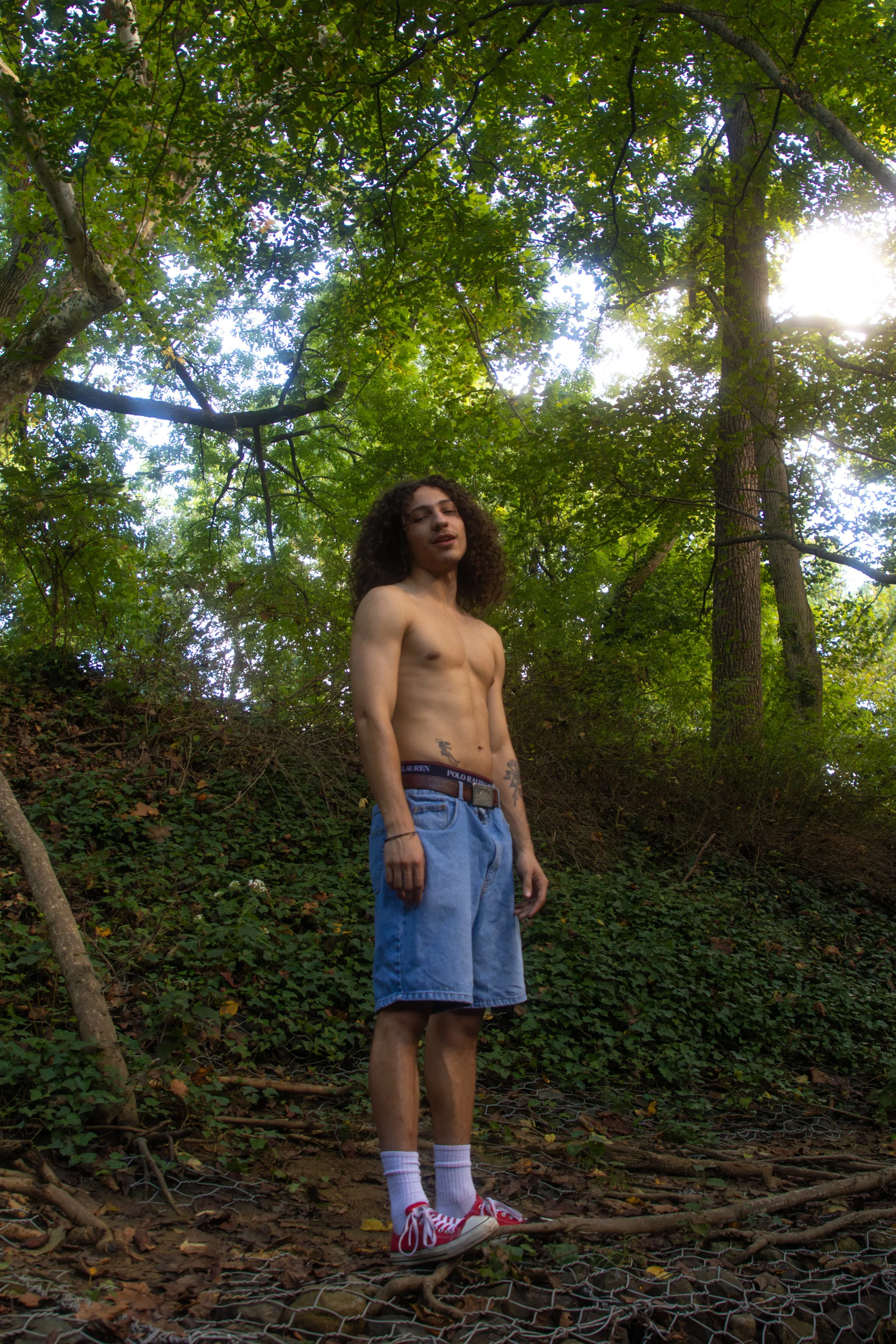 A shirtless man with curly hair standing outdoors amidst trees and greenery, wearing denim shorts, red sneakers, and white socks.
