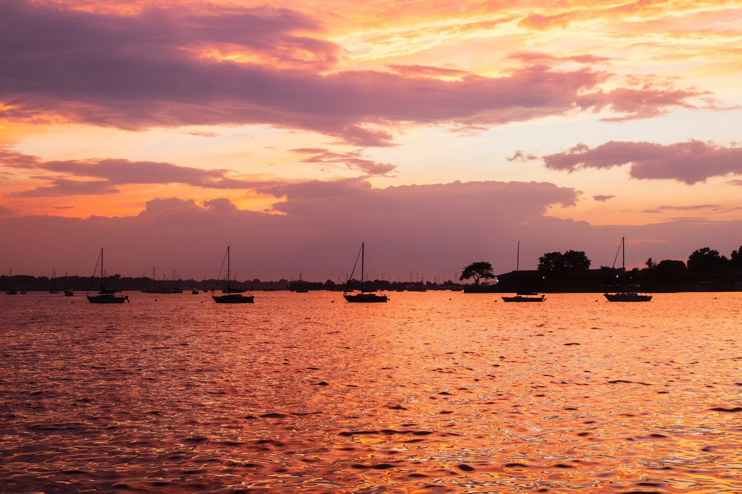 Sunset over a body of water with sailboats anchored, colorful sky with clouds, and silhouettes of trees and buildings in the distance.