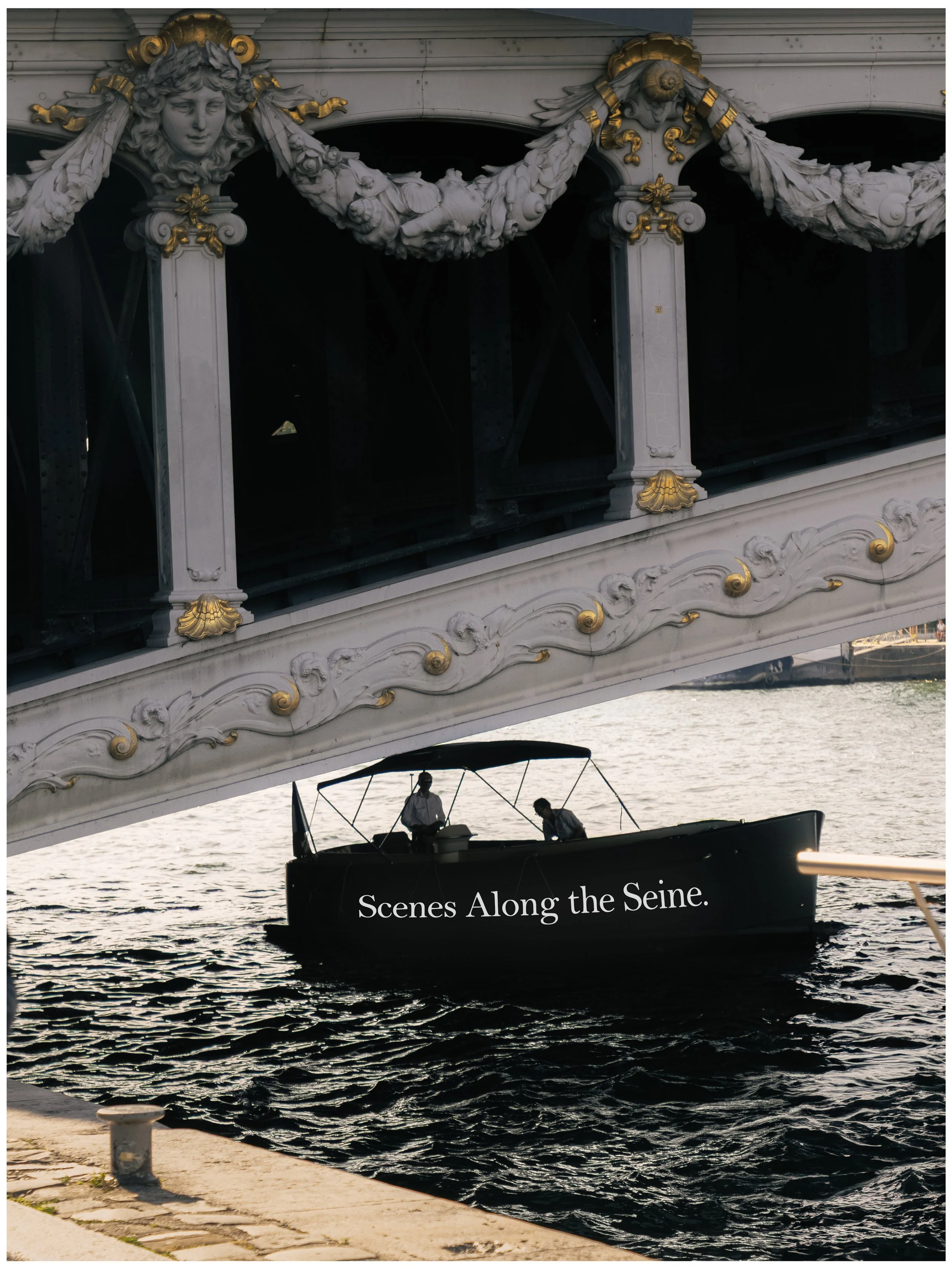 A boat on the water under a bridge with ornate decoration. The boat has the words "Scenes Along the Seine." written on its side, and two people can be seen on it.
