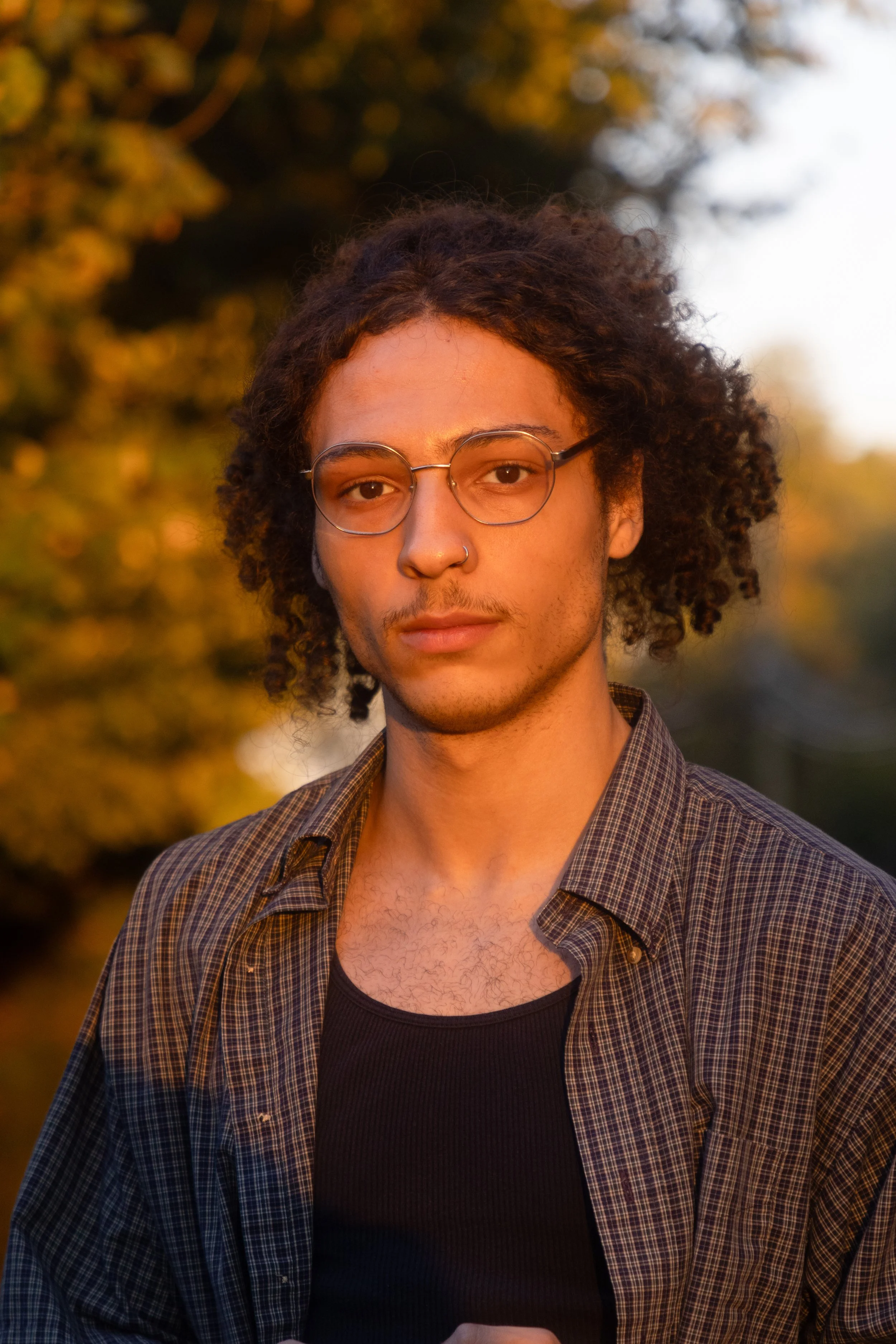 A young man with curly hair, glasses, and a nose ring looking at the camera outdoors during sunset.