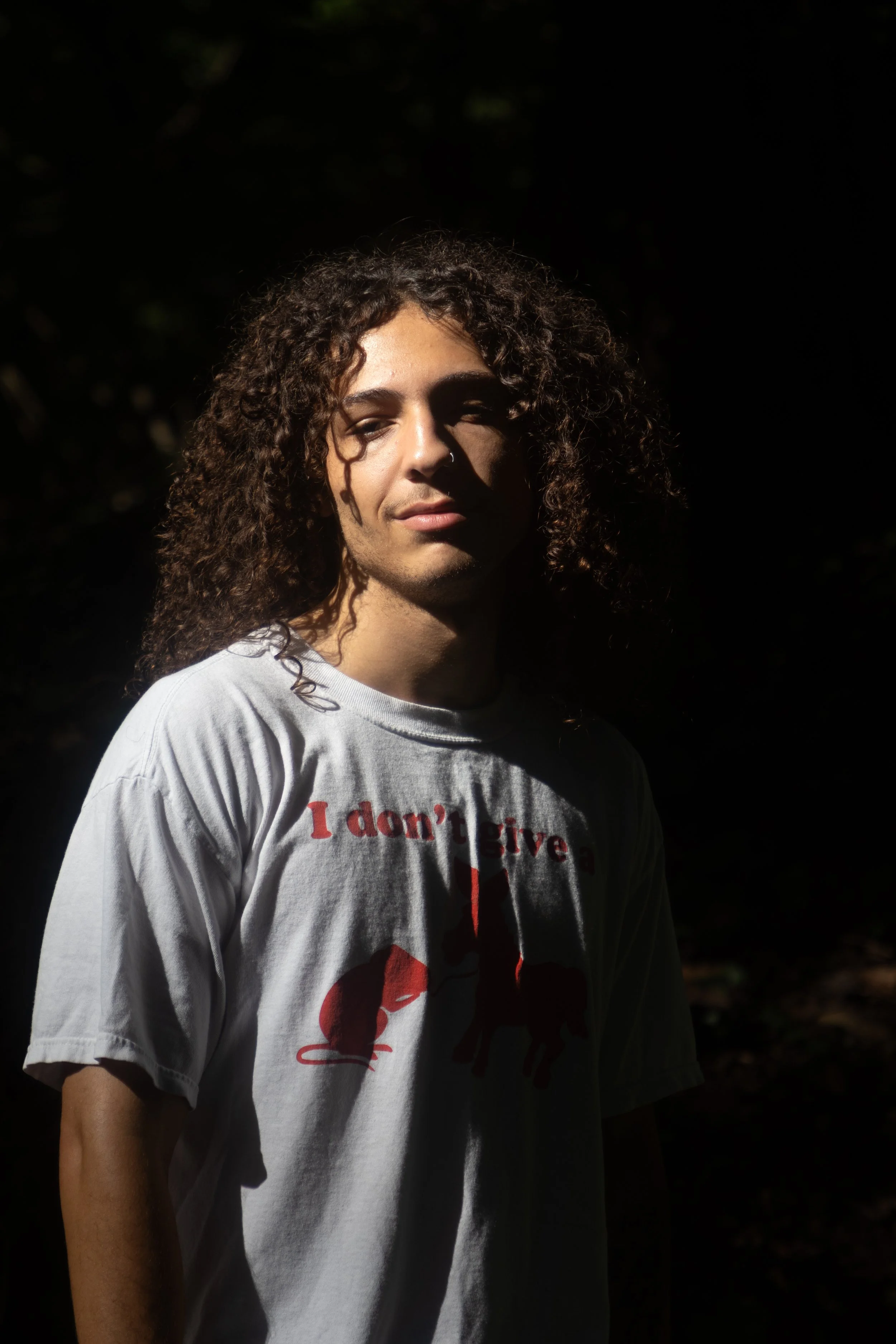 Person with long curly hair and light skin, wearing a white T-shirt with red text and images, standing outdoors in partial shadow with a dark background.