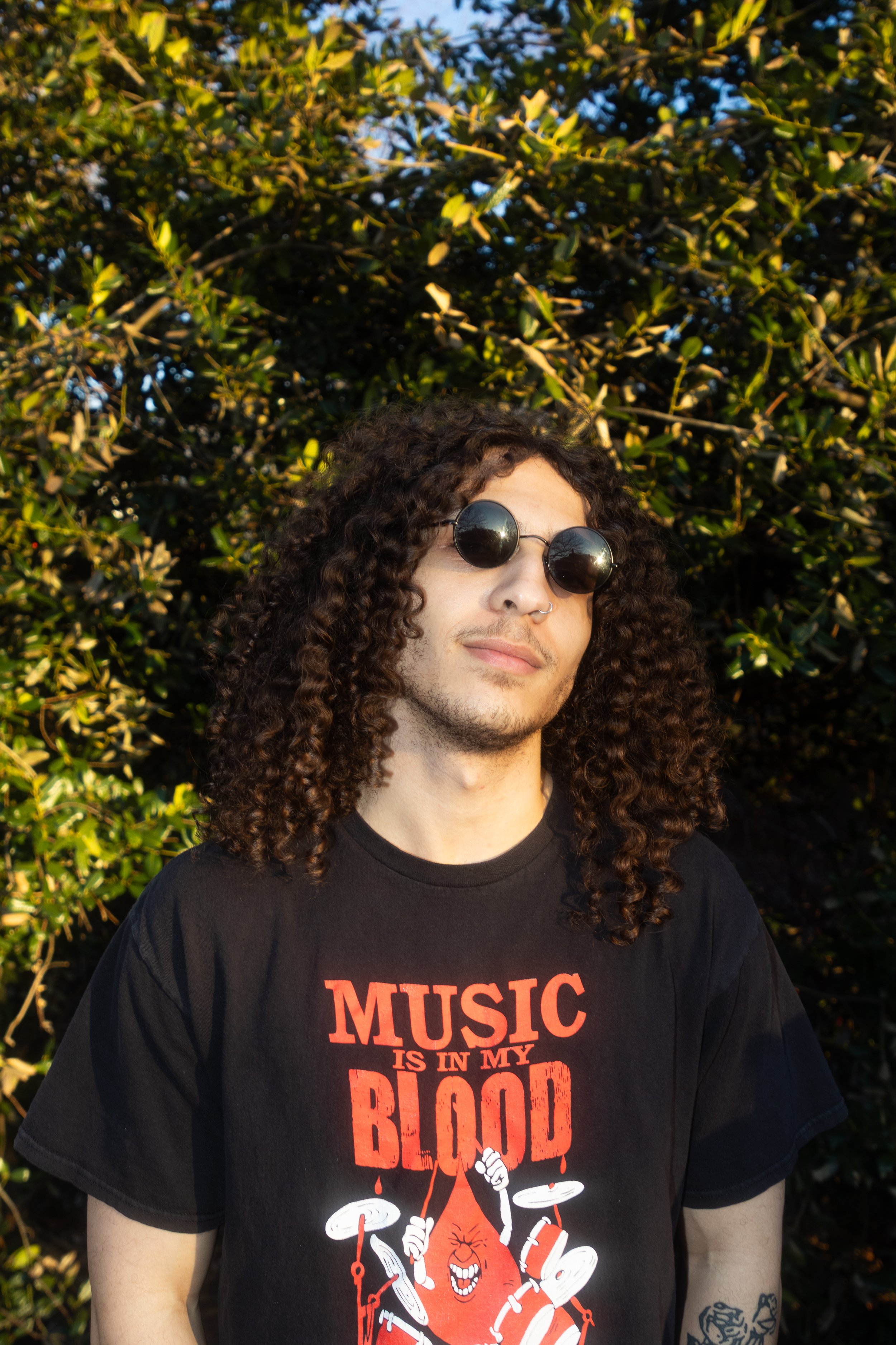 A young man with curly hair wearing sunglasses and a black T-shirt with a graphic and text, standing outdoors in front of green bushes on a sunny day.