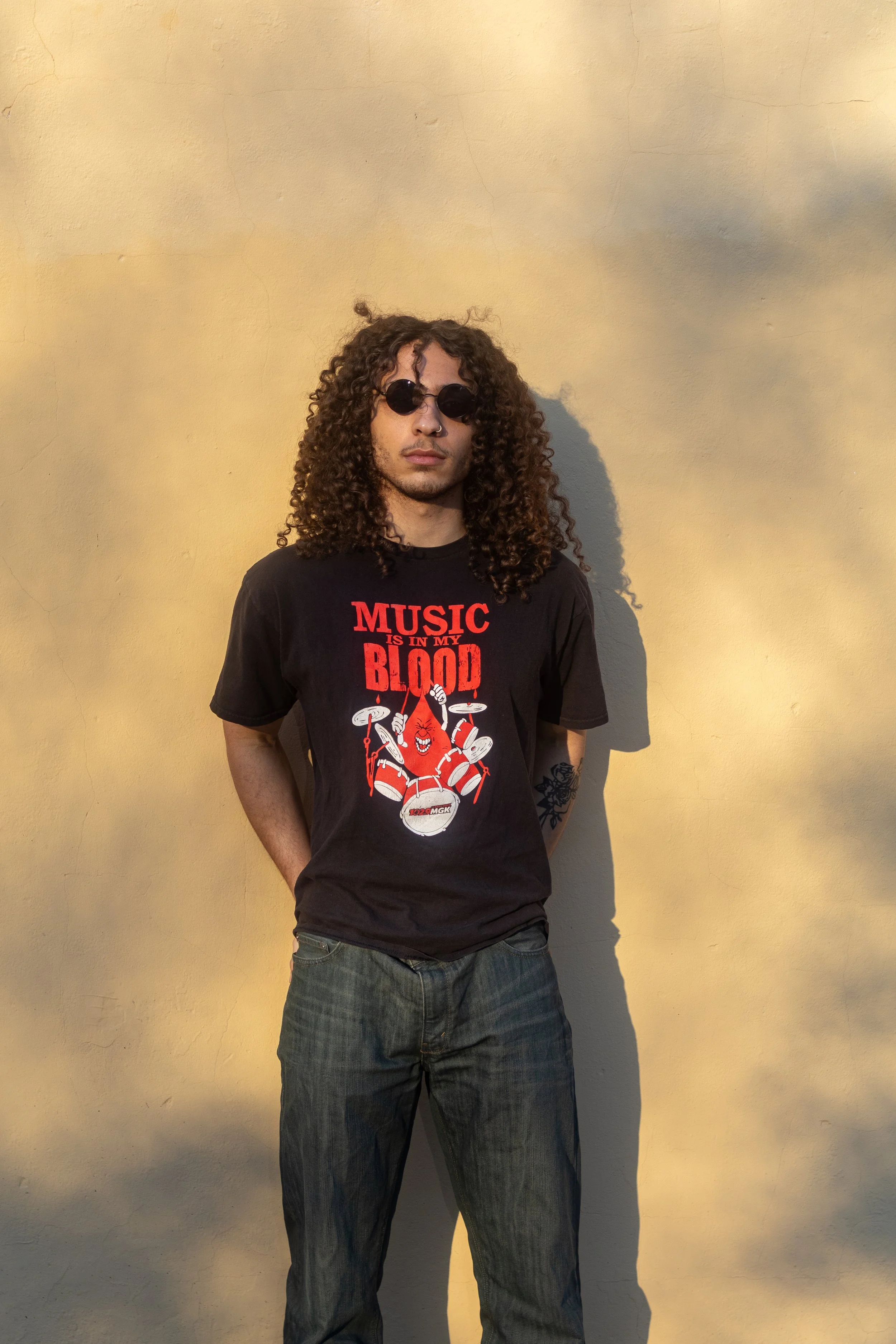 A young man with curly hair wearing sunglasses, a black graphic T-shirt that says 'Music is in my blood' with a red blood drop and a cartoon drummer, and jeans, standing against a yellow wall with shadows.