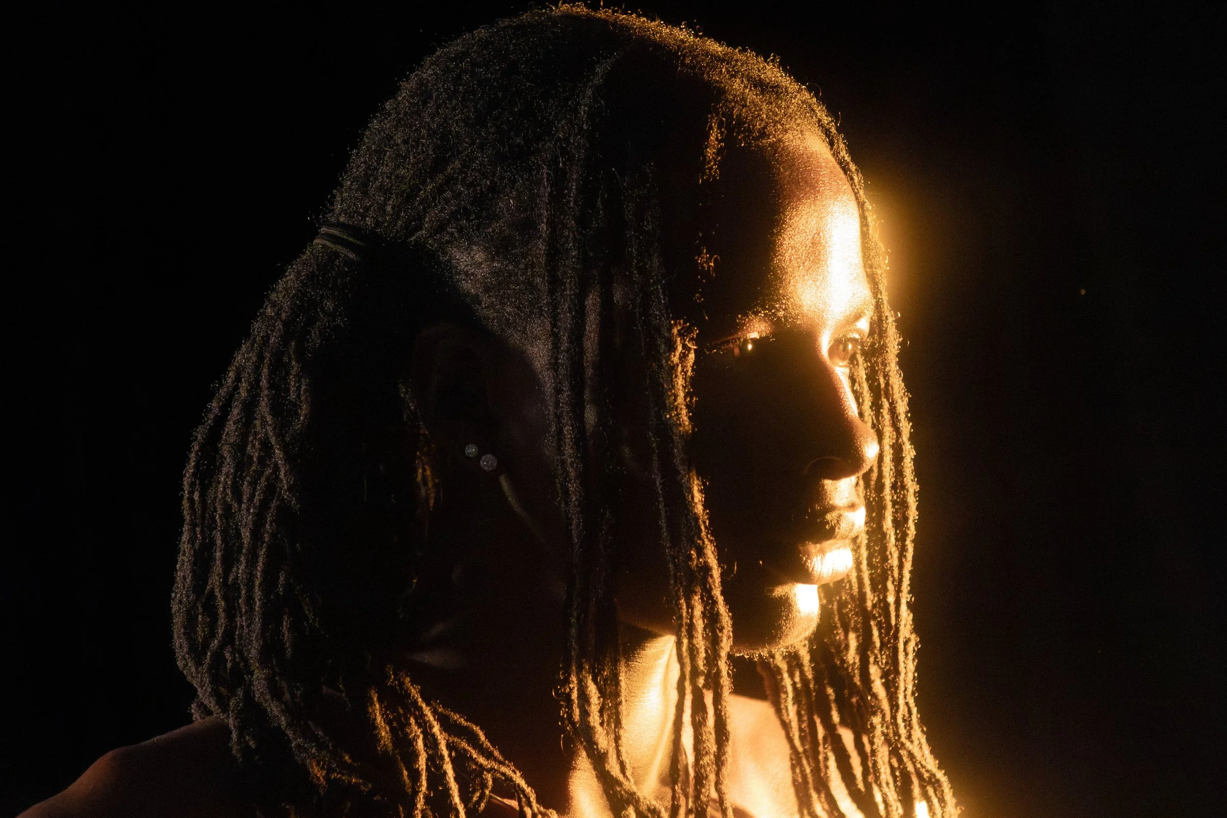 Profile of a woman with dreadlocks lit by warm, golden light against a dark background.