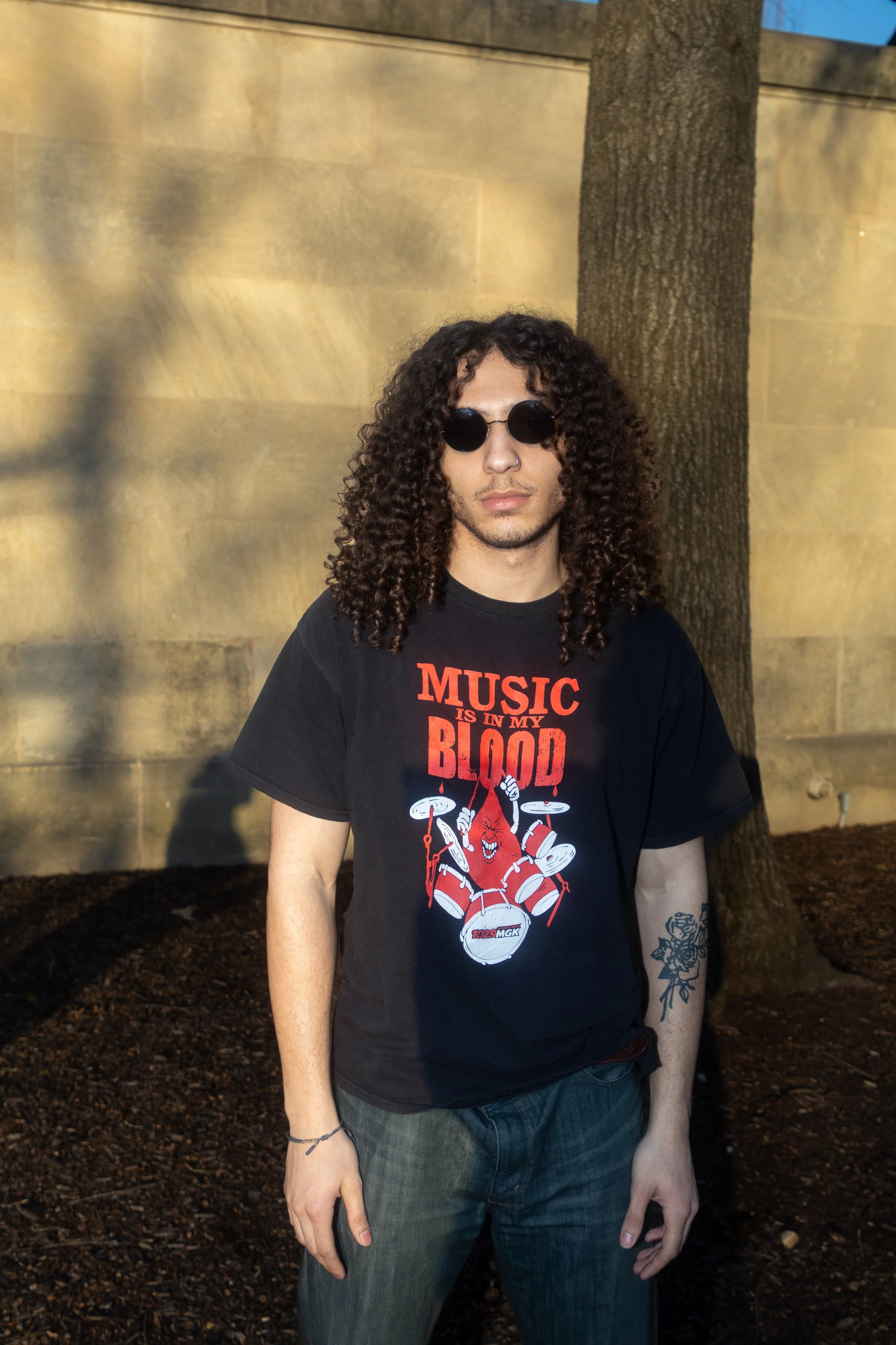 A young man with long curly hair, wearing round black sunglasses, a black T-shirt with a graphic of a red cartoon character playing drums and the text 'Music is in my blood,' and blue jeans, stands outdoors next to a large tree and a concrete wall in