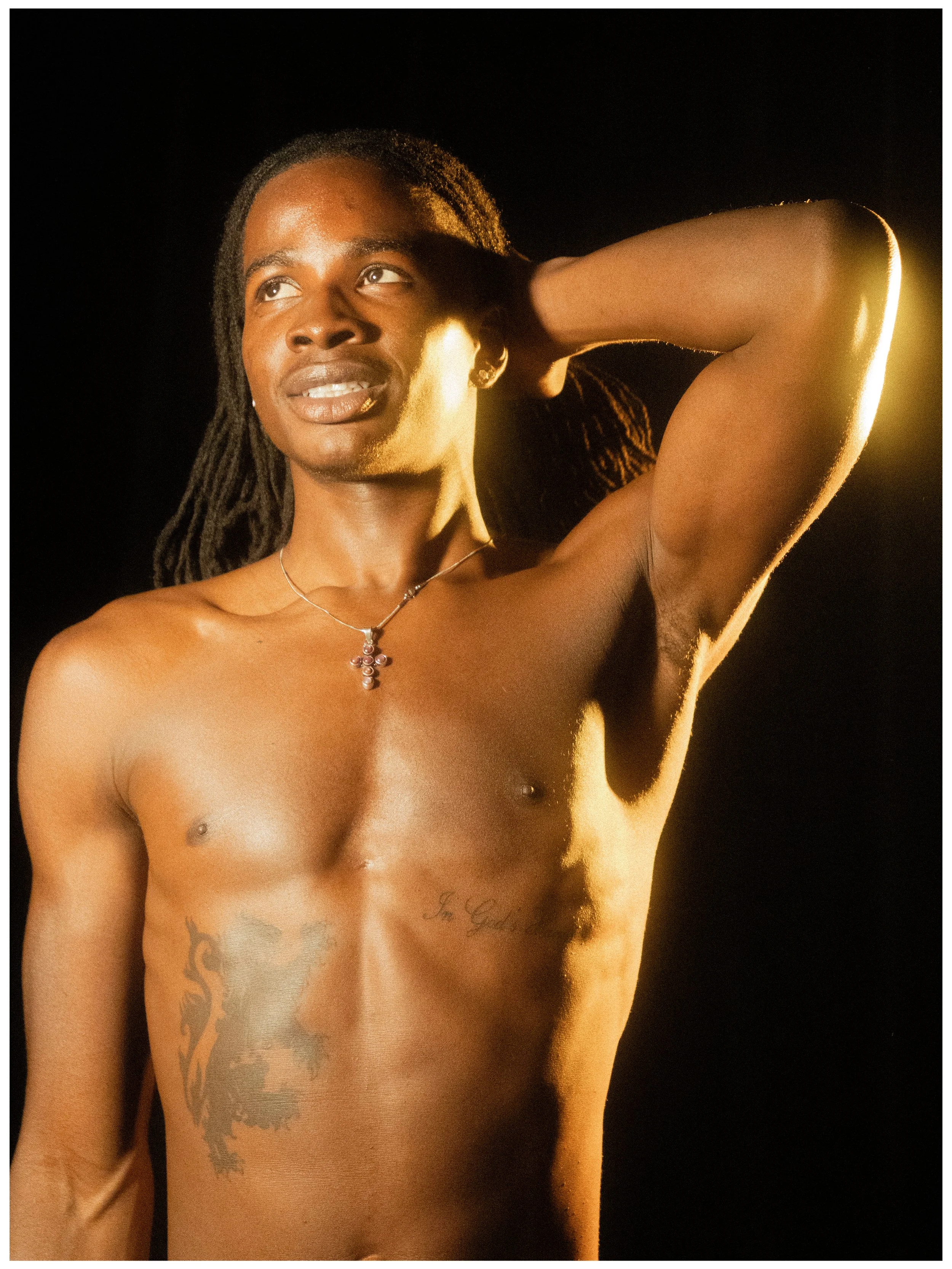 A topless man with dreadlocks and tattoos on his chest, wearing a necklace, posing with one arm raised behind his head against a dark background.