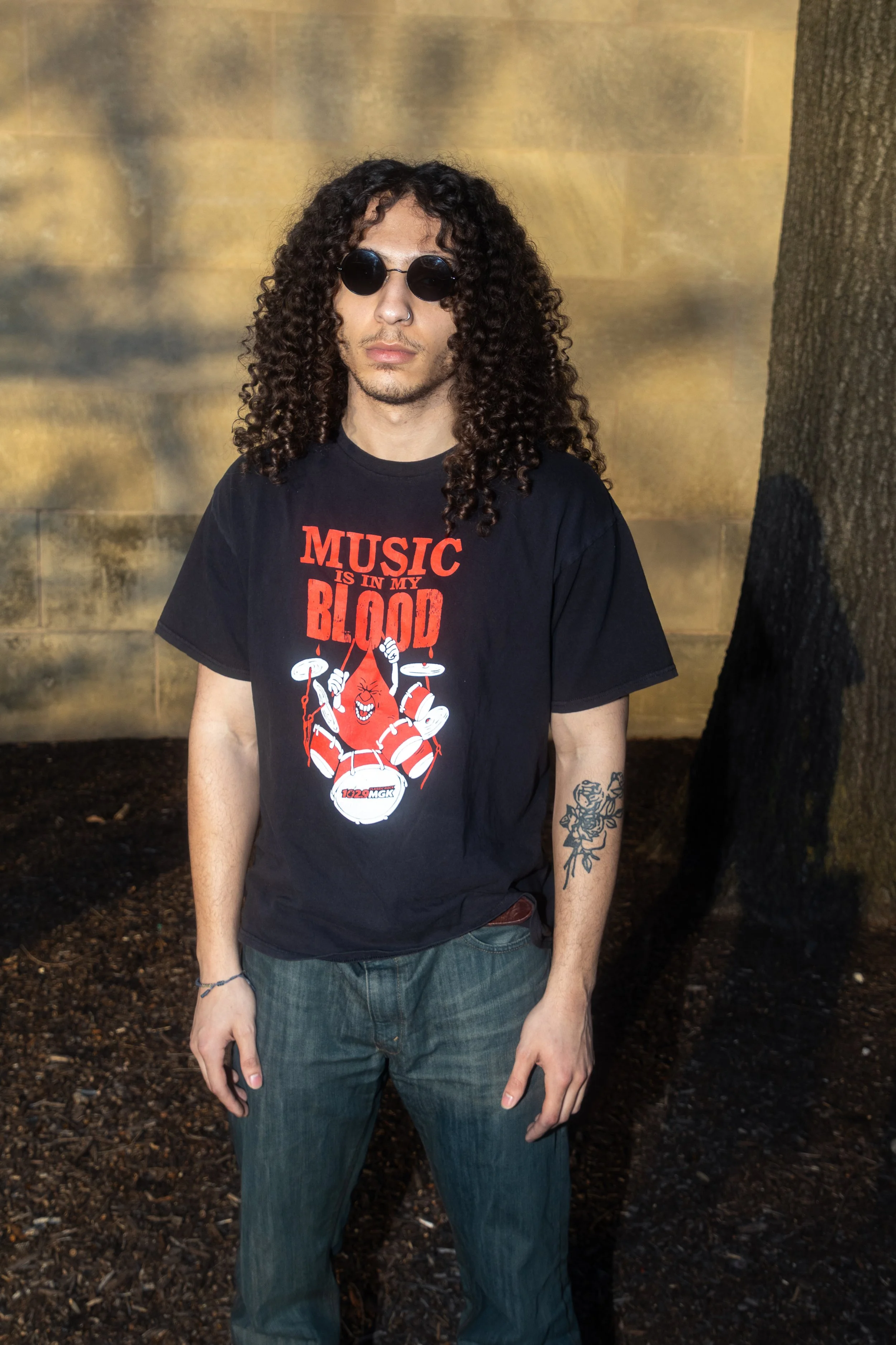 A man with curly hair wearing sunglasses, a black T-shirt with red and white graphics, and blue jeans, standing outdoors next to a tree and a stone wall.