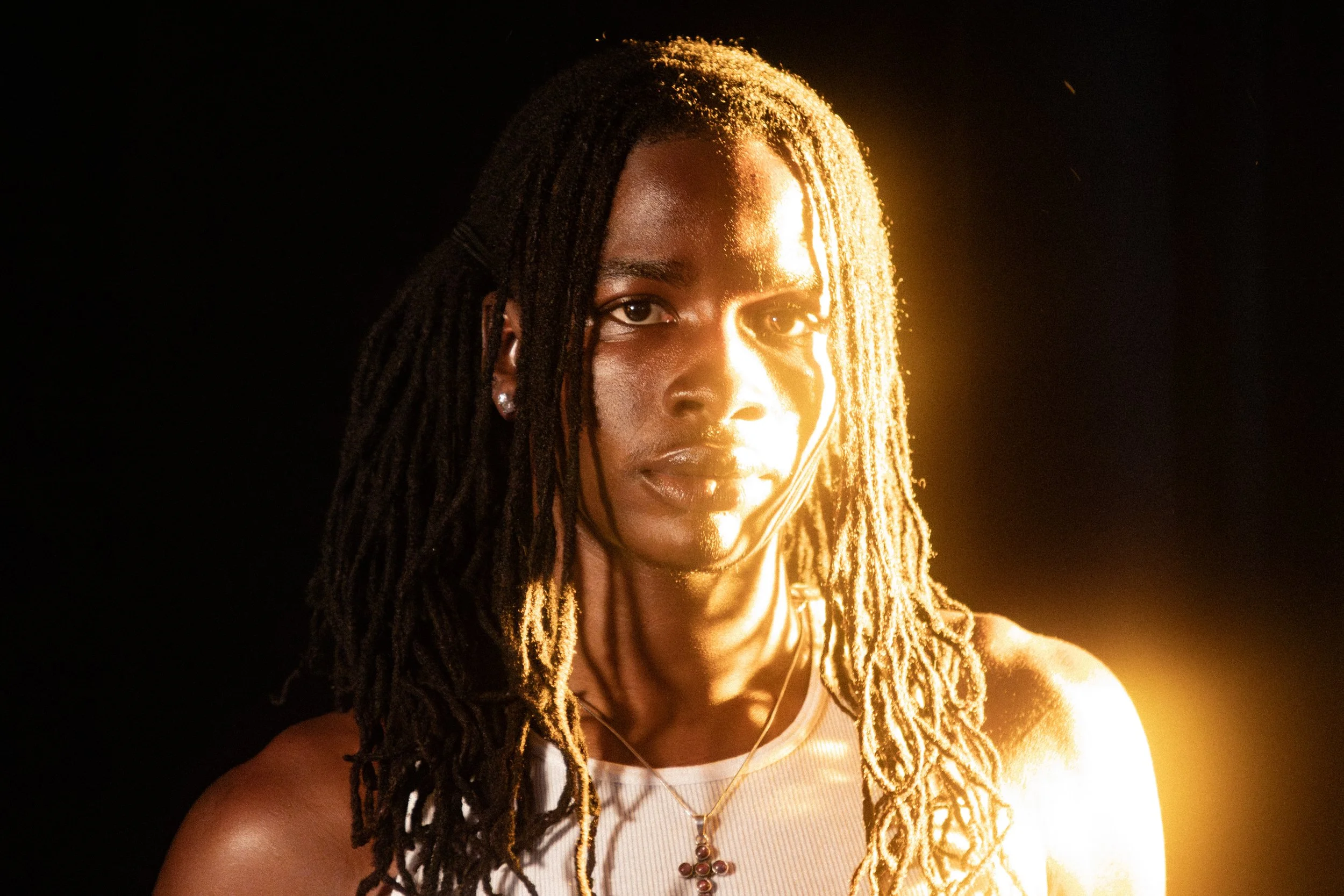 A woman with dreadlocks and earrings, wearing a white top and a cross necklace, illuminated by a warm, golden light against a dark background.