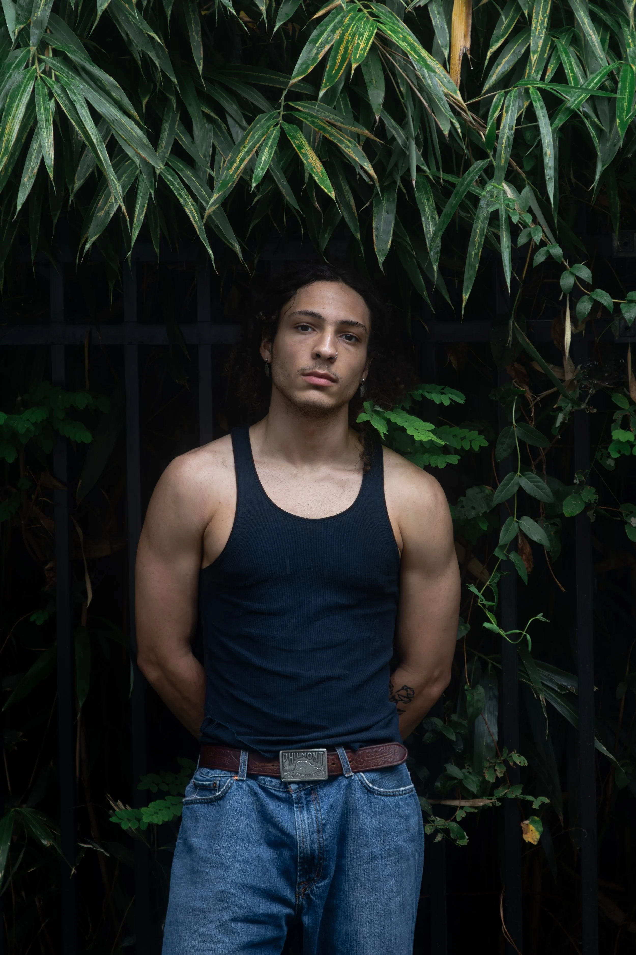 A young man with dark curly hair and light skin, wearing a navy tank top and blue jeans, standing in front of green leafy plants and black metal fence.