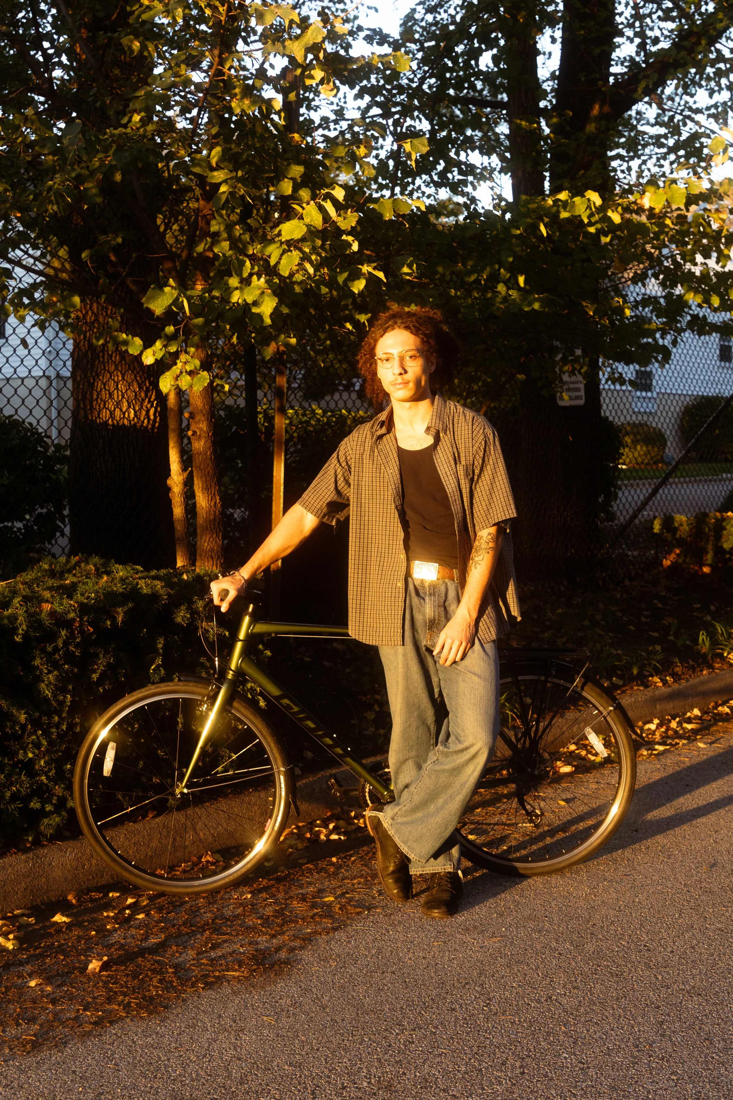 A person with curly hair and glasses standing next to a bicycle on a sidewalk, illuminated by warm street lighting in the evening, with trees and a fence in the background.