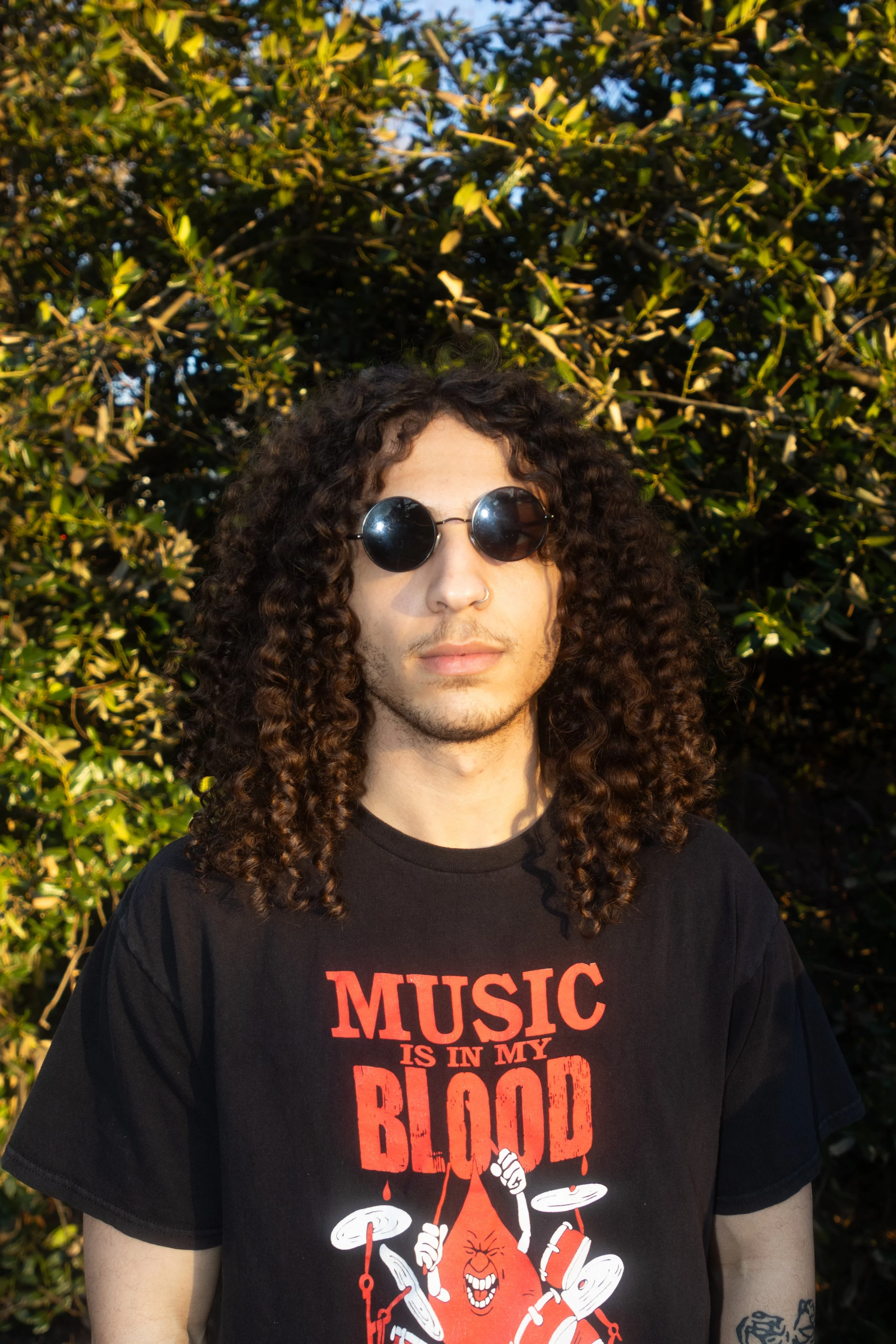 A young man with curly hair and sunglasses standing outdoors in front of dense green foliage, wearing a black T-shirt with a red and white graphic design and text that reads 'Music is in my blood'.