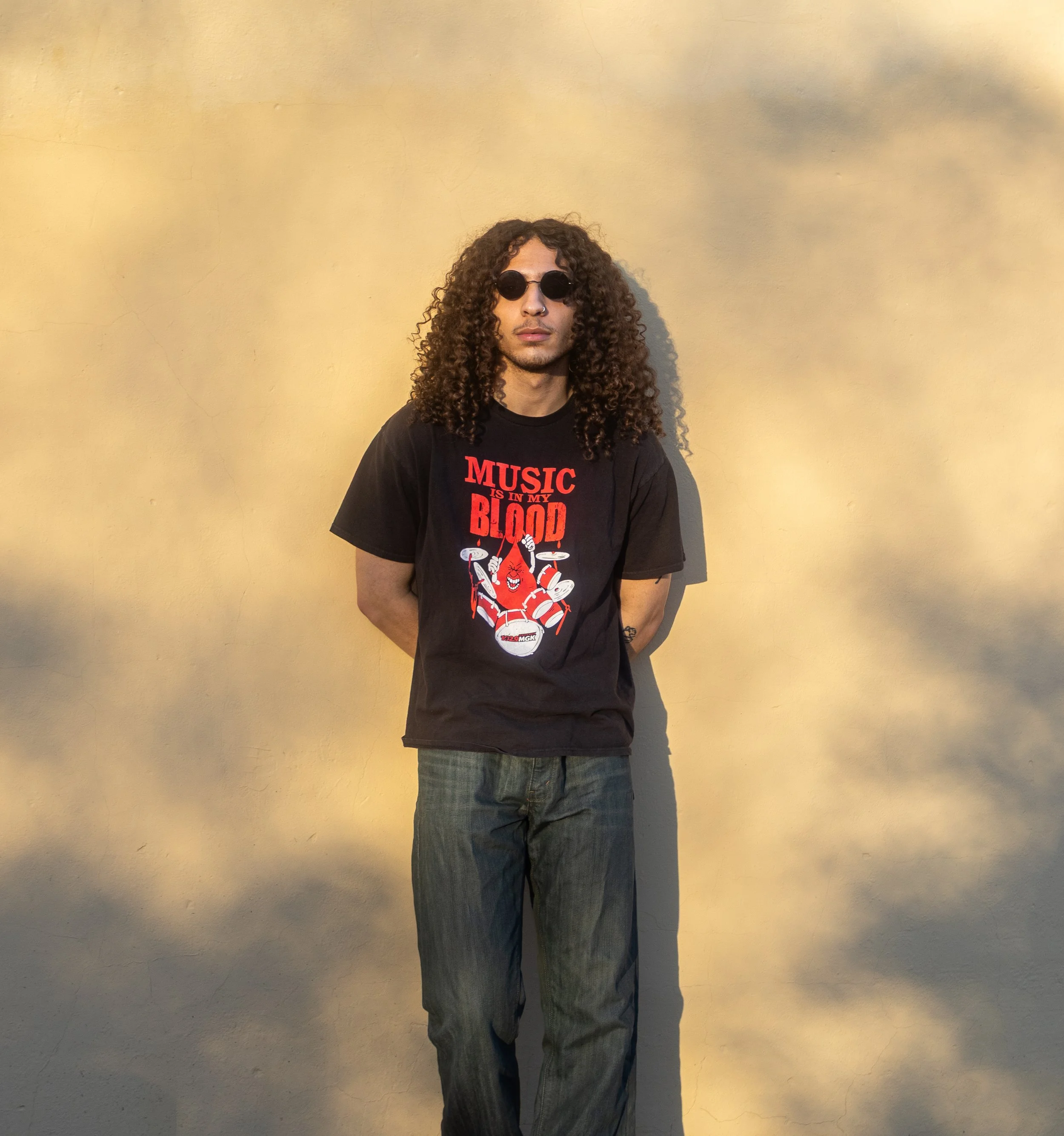 A young man with long curly hair wearing black sunglasses, a black graphic T-shirt with red text that reads 'Music is in my blood' and a drum set illustration, and dark jeans, standing against a beige wall with shadows cast on it.