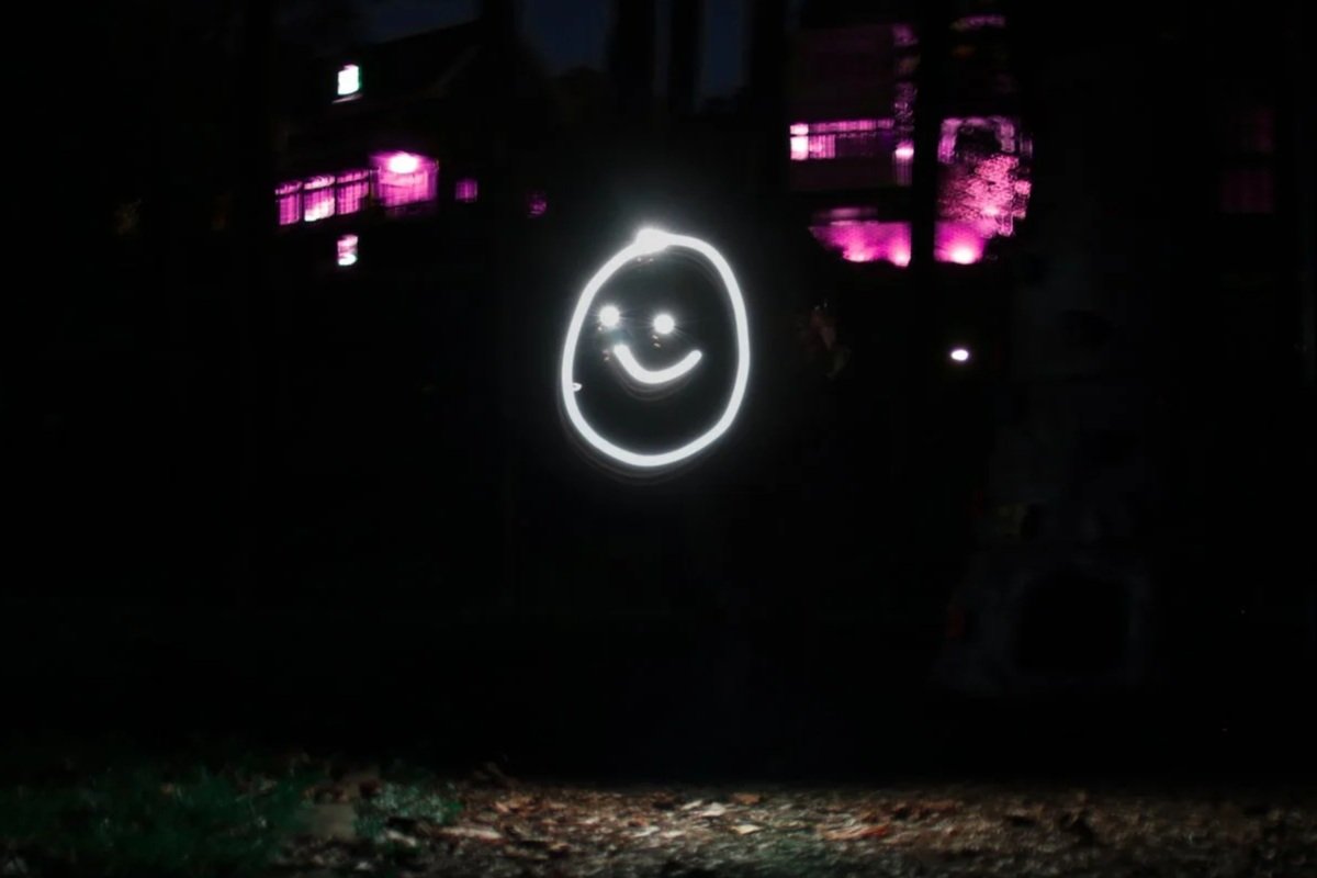 Person holding a light-up sign with a smiling face drawn in white light, in a dark outdoor setting with buildings illuminated in pink in the background. Light painting.