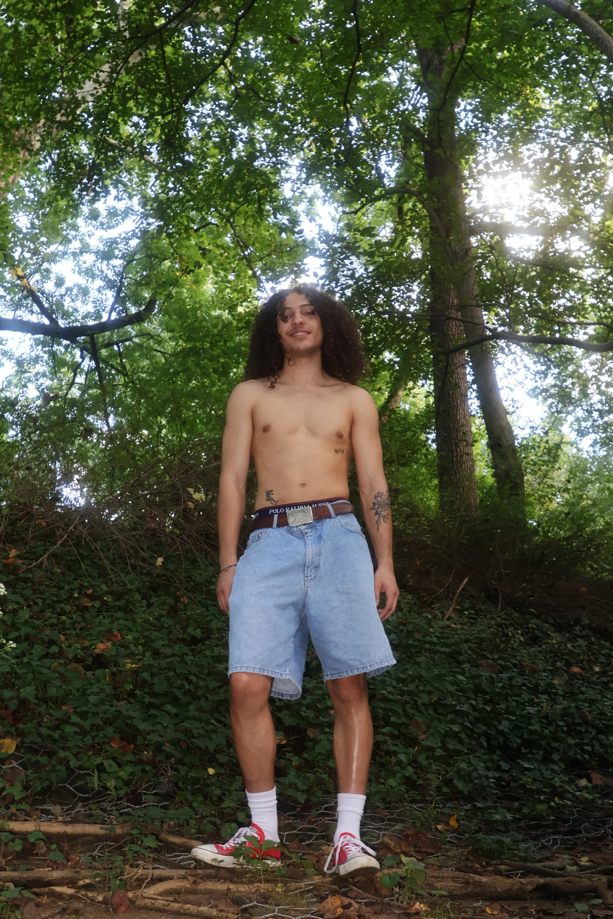 A shirtless young man with long curly hair standing outdoors in a lush green forest, wearing light denim shorts, white socks, and red and white sneakers, looking at the camera and smiling.