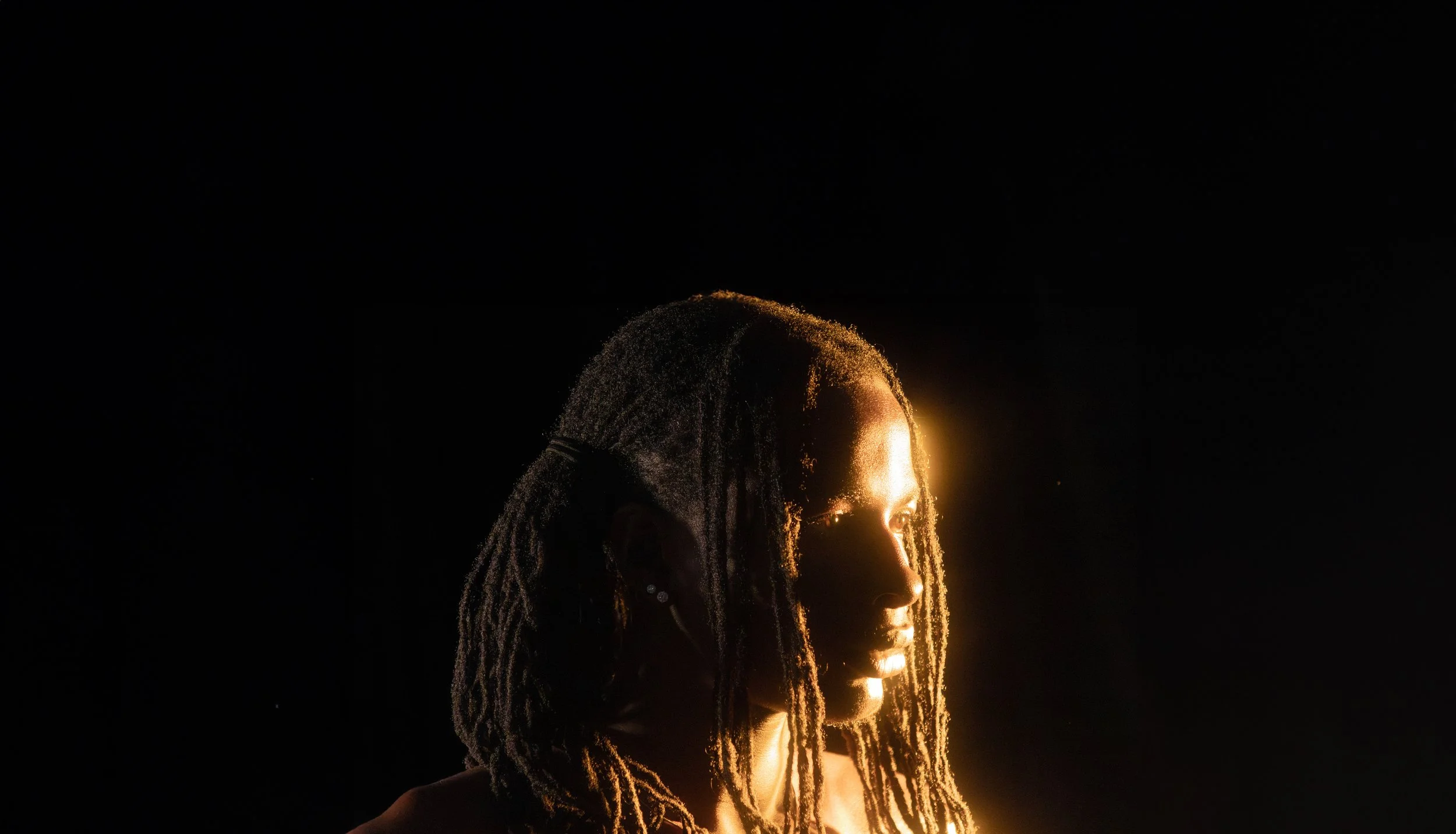 Side profile of a woman with dreadlocks illuminated by a warm light, against a black background.