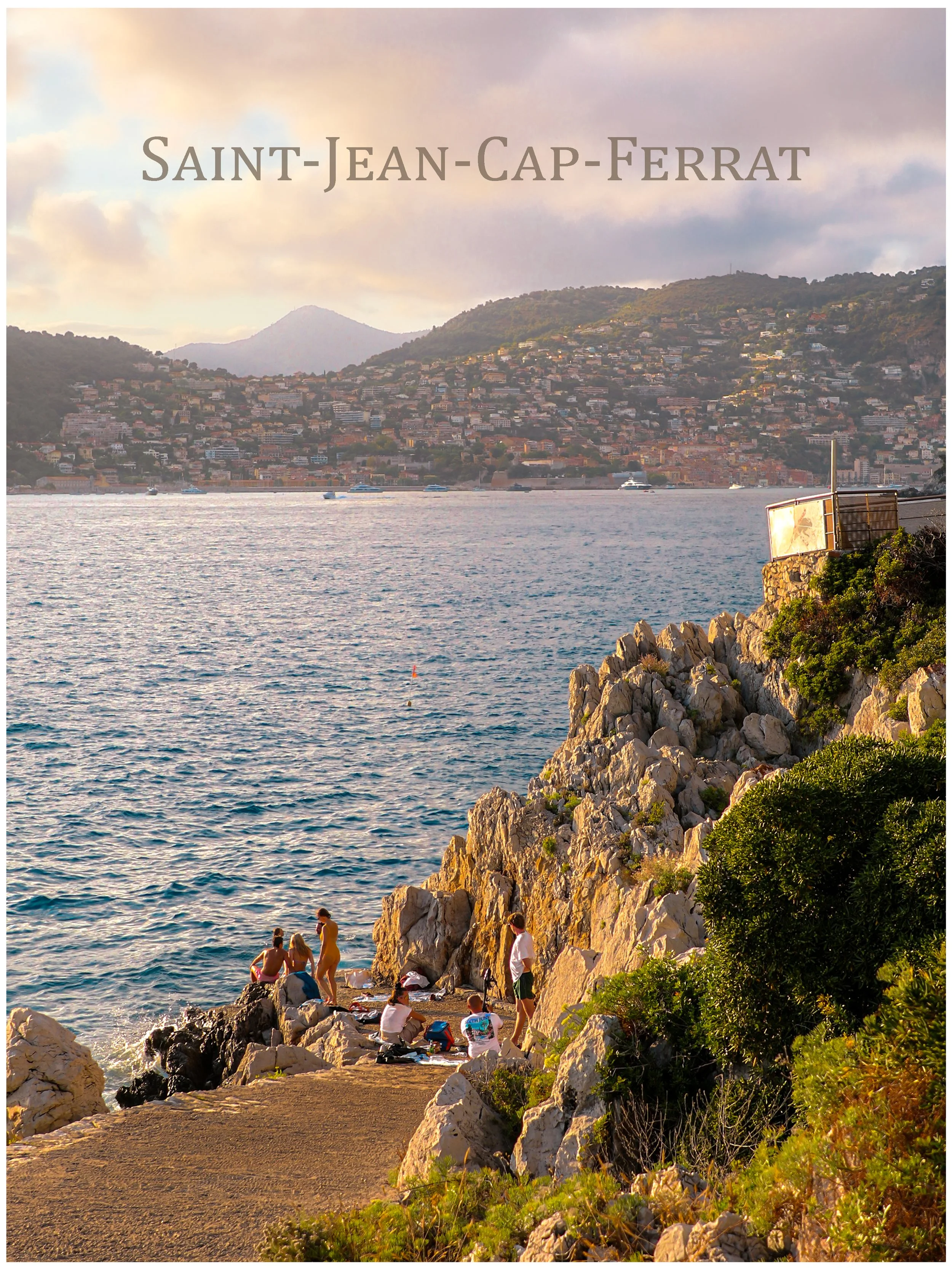 People sitting and standing on rocks and a path by the water's edge near a rocky coastline with greenery, with a city and hillside in the background under a cloudy sky.