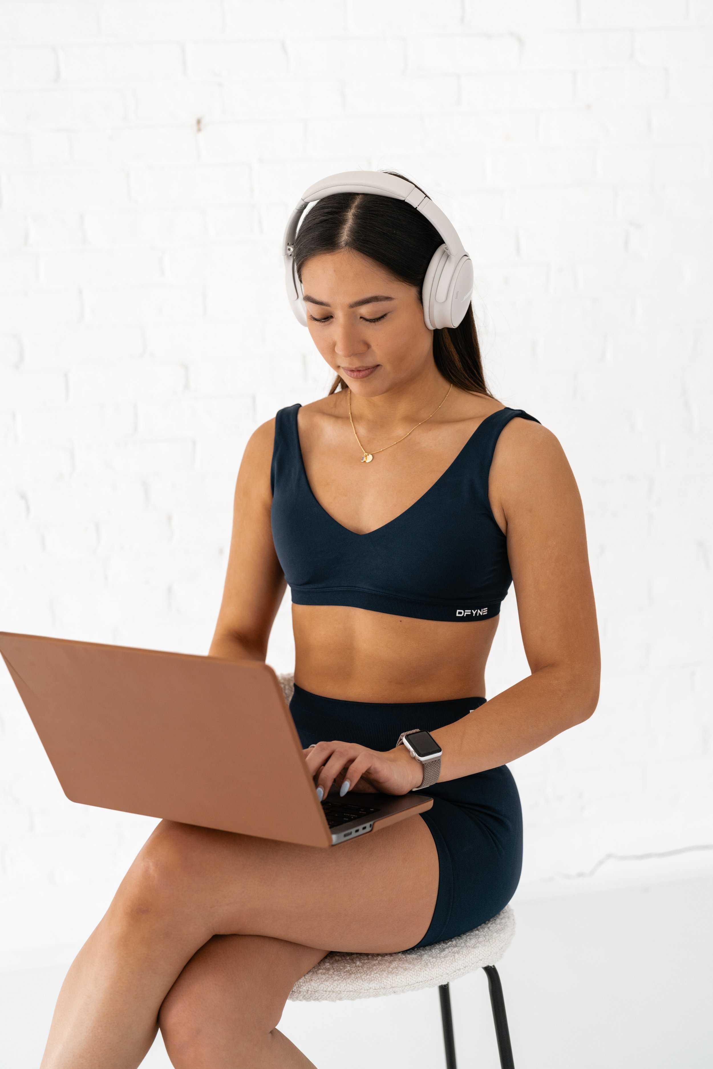 A woman wearing a navy sports bra and shorts sitting on a stool, using a laptop, wearing headphones, and a smartwatch, with a white brick wall in the background.