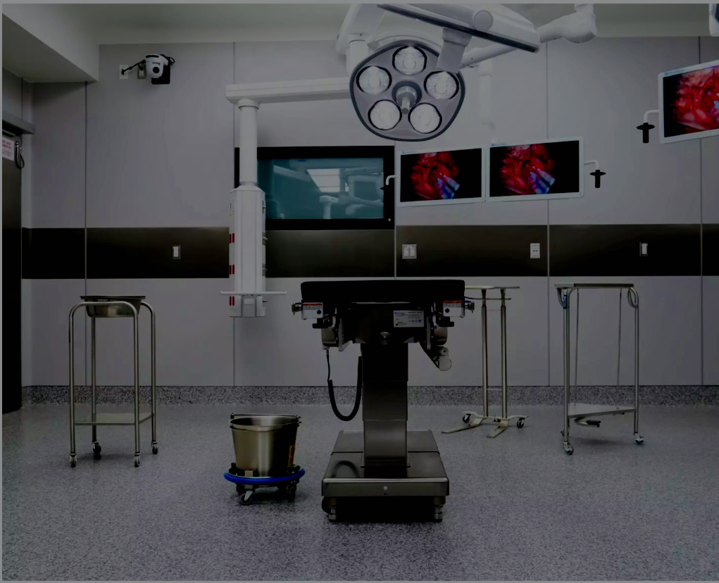 Empty operating room with surgical table, overhead surgical lights, monitors on the wall, and medical equipment.