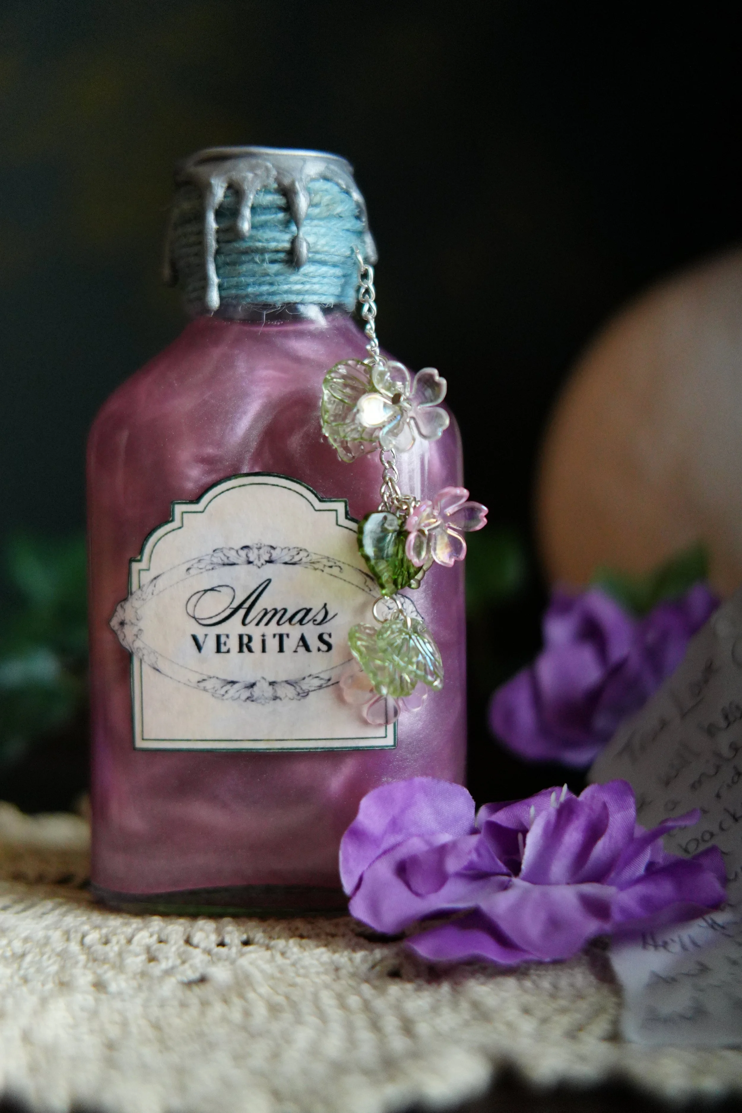 A decorative pink potion bottle labeled “Amas Veritas,” sealed with a blue wax-style cap and adorned with floral charms, surrounded by purple flowers on a textured surface.
