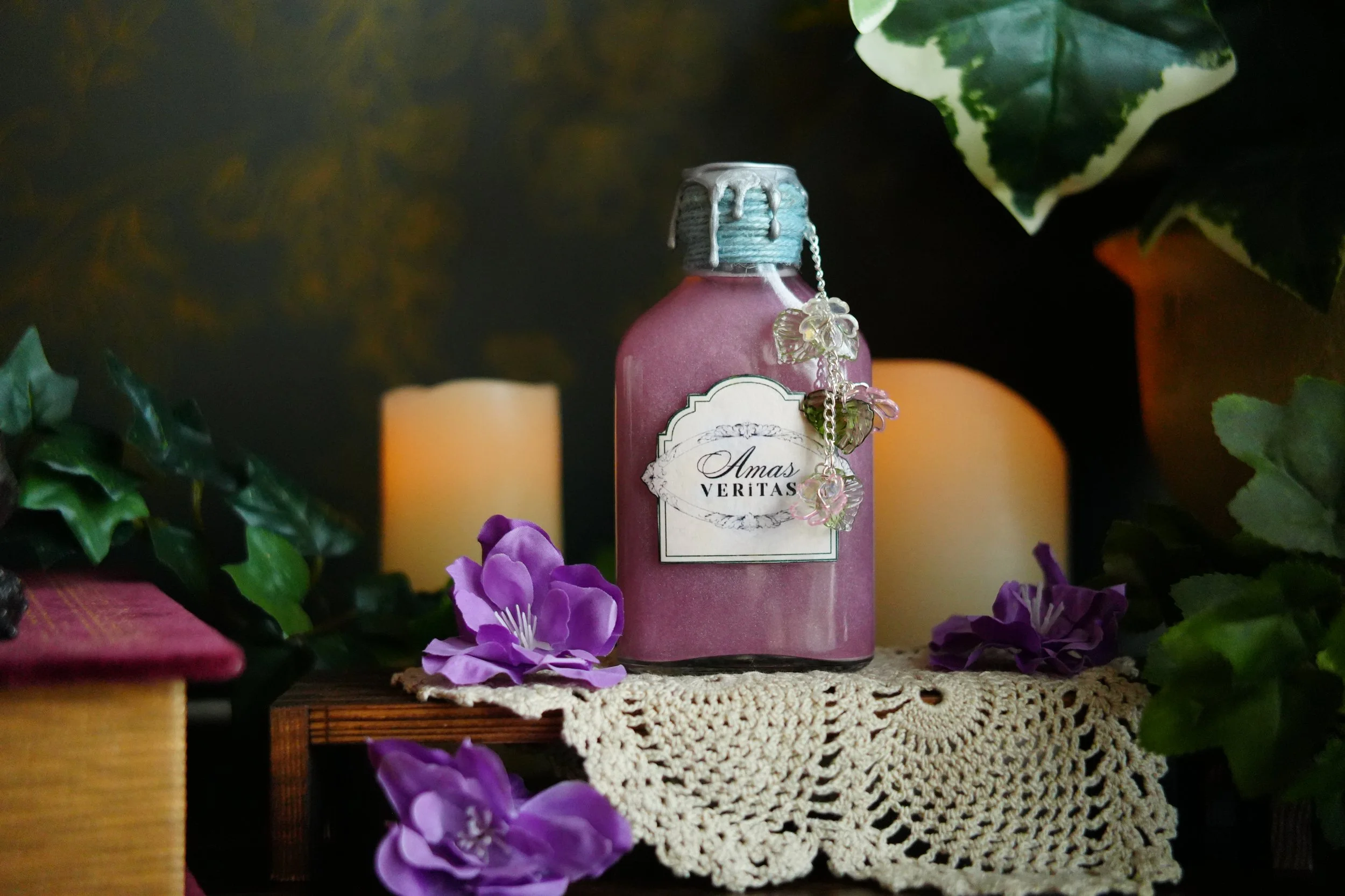 A decorative pink potion bottle labeled “Amas Veritas,” sealed with a blue wax-style cap and floral charms, displayed among purple flowers, candles, greenery, and a vintage crochet cloth.