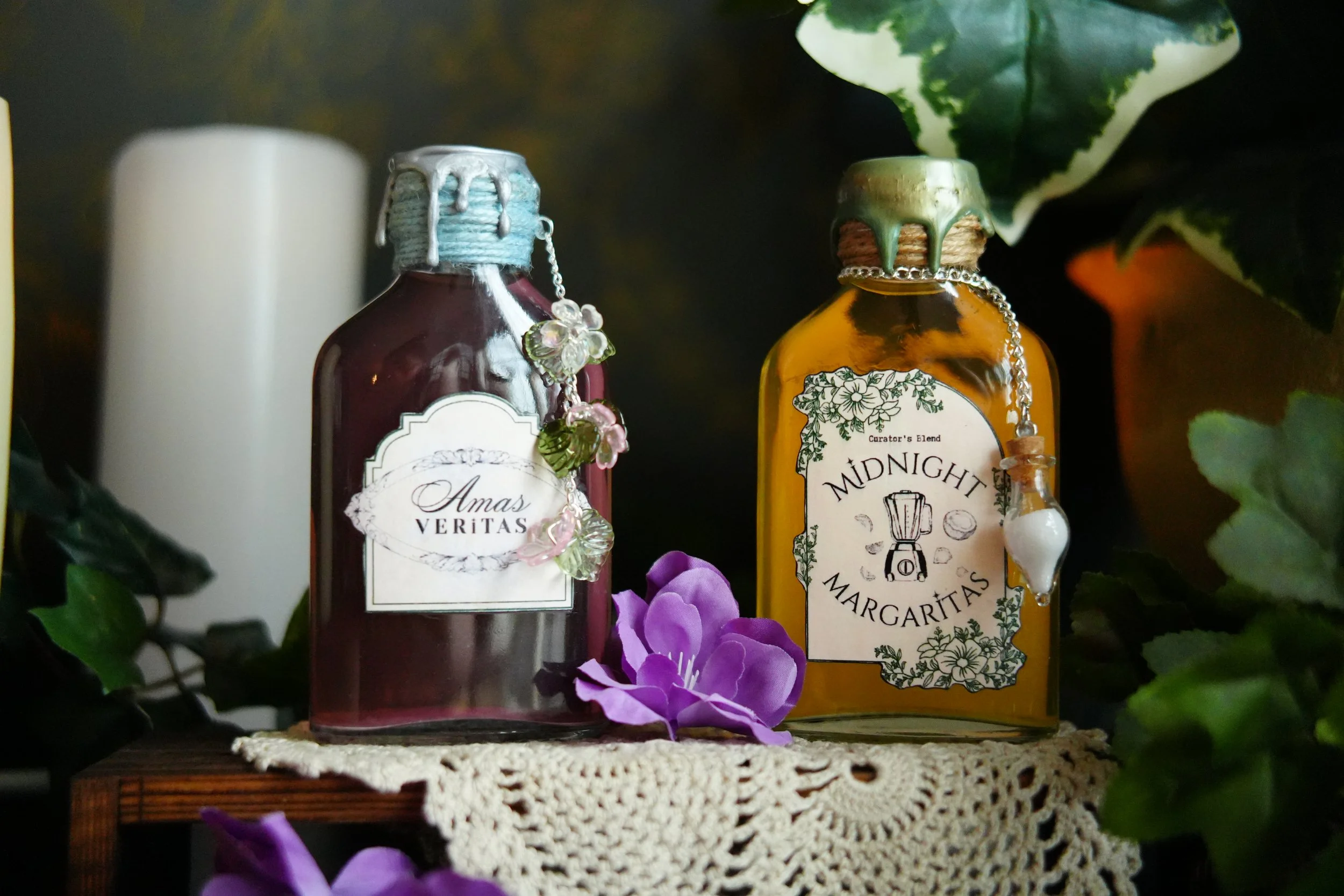 A pink “Amas Veritas” potion bottle and golden “Midnight Margaritas” potion bottle with wax-sealed caps and floral charms, displayed among purple flowers, greenery, and a vintage crochet cloth.