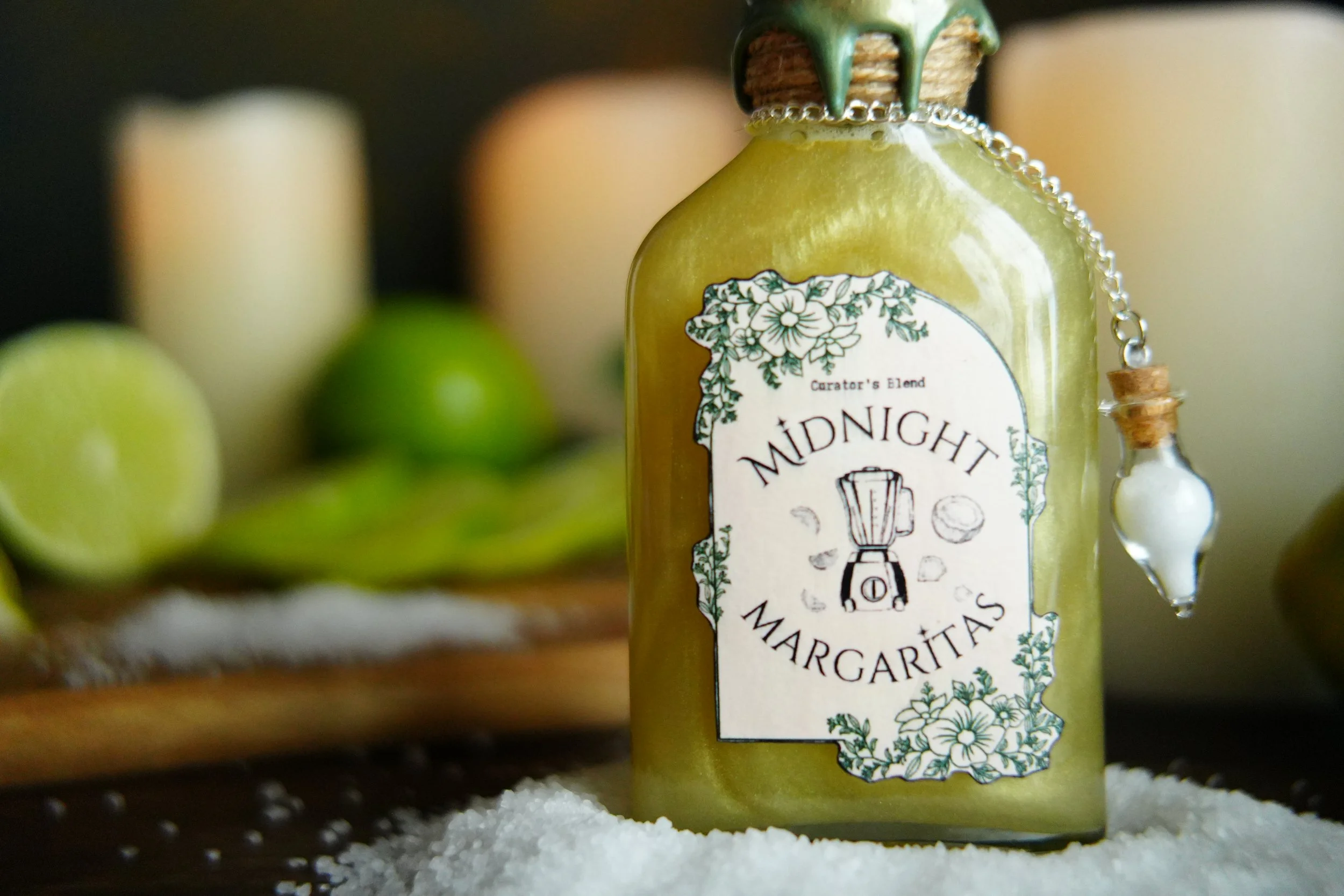 Close-up of a golden “Midnight Margaritas” potion bottle with a wax-sealed cap and dangling charm, resting in salt with limes and candles blurred in the background.