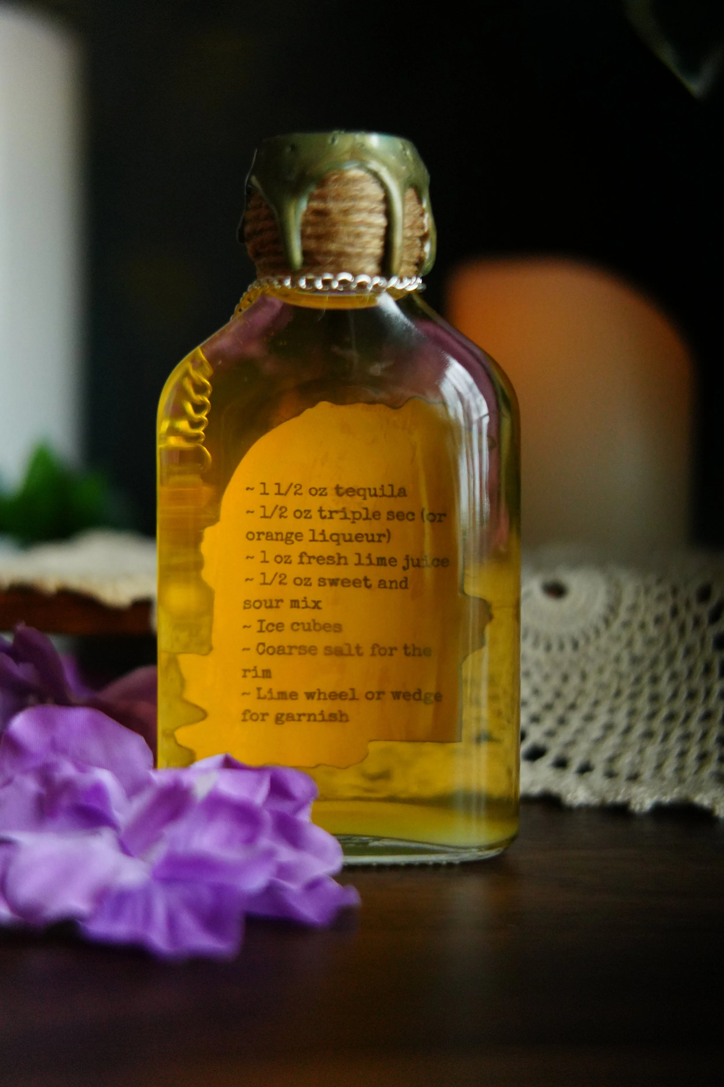A golden “Midnight Margaritas” potion bottle with a wax-sealed cap, displaying a typewriter-style margarita recipe, surrounded by purple flowers and a vintage crochet cloth.