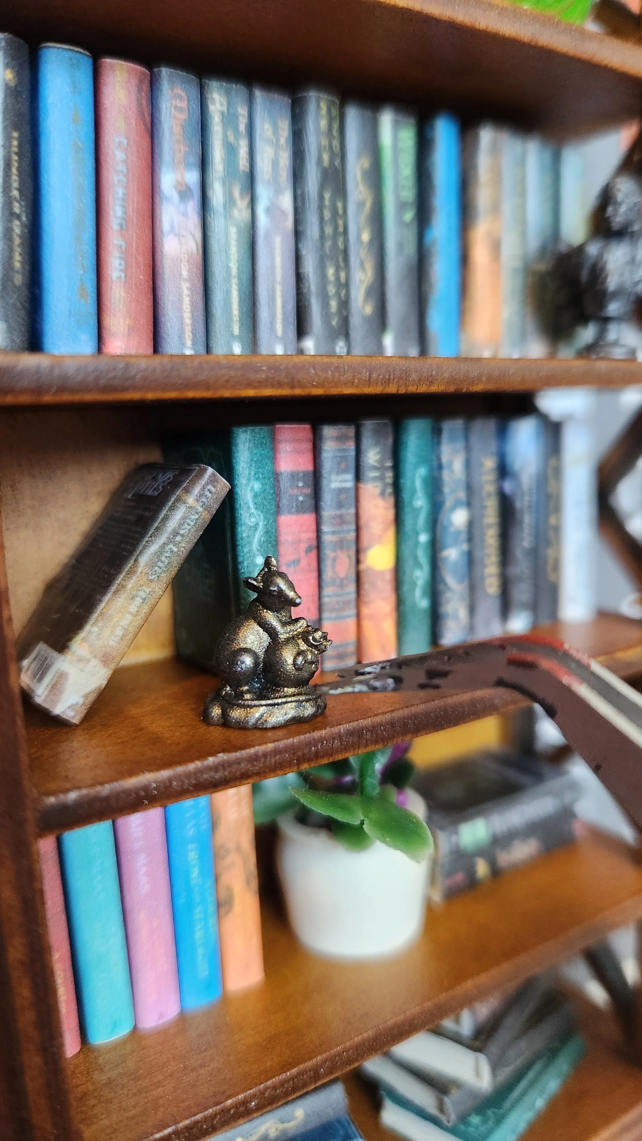 A miniature wooden bookshelf with colorful miniature books, a small mouse figurine, a tiny potted plant, and other decorative items.
