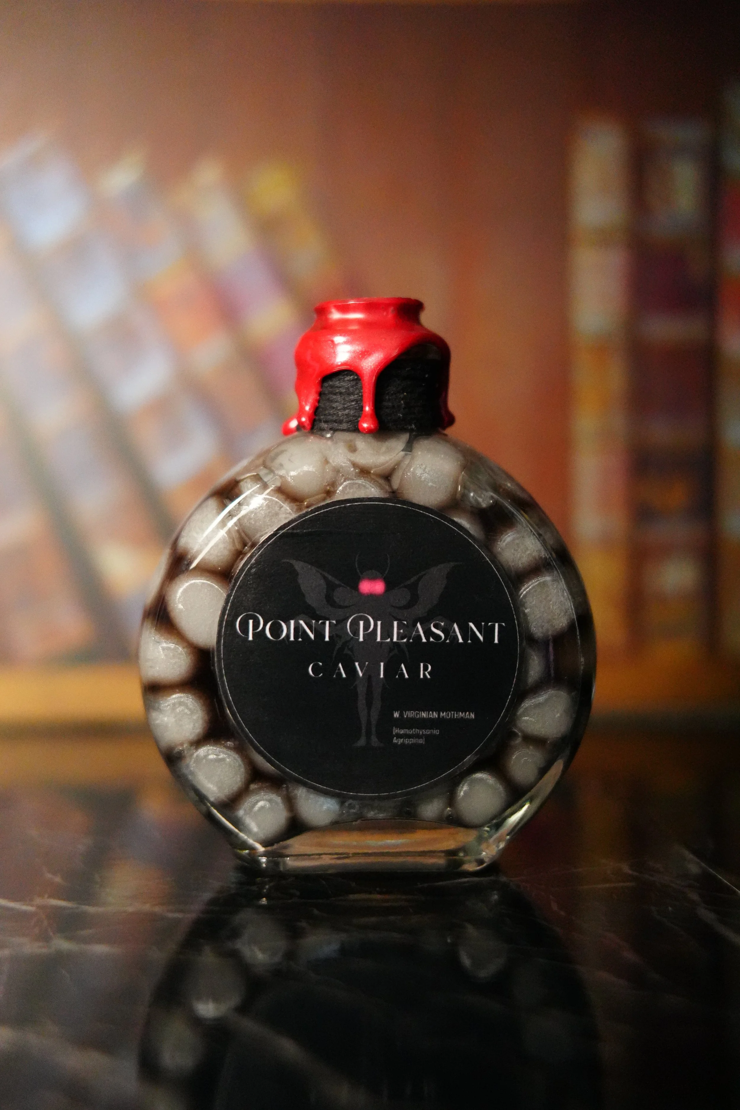 Round glass bottle labeled "Point Pleasant Caviar" filled with shimmering gray beads, topped with dripping red wax against a softly blurred library background.